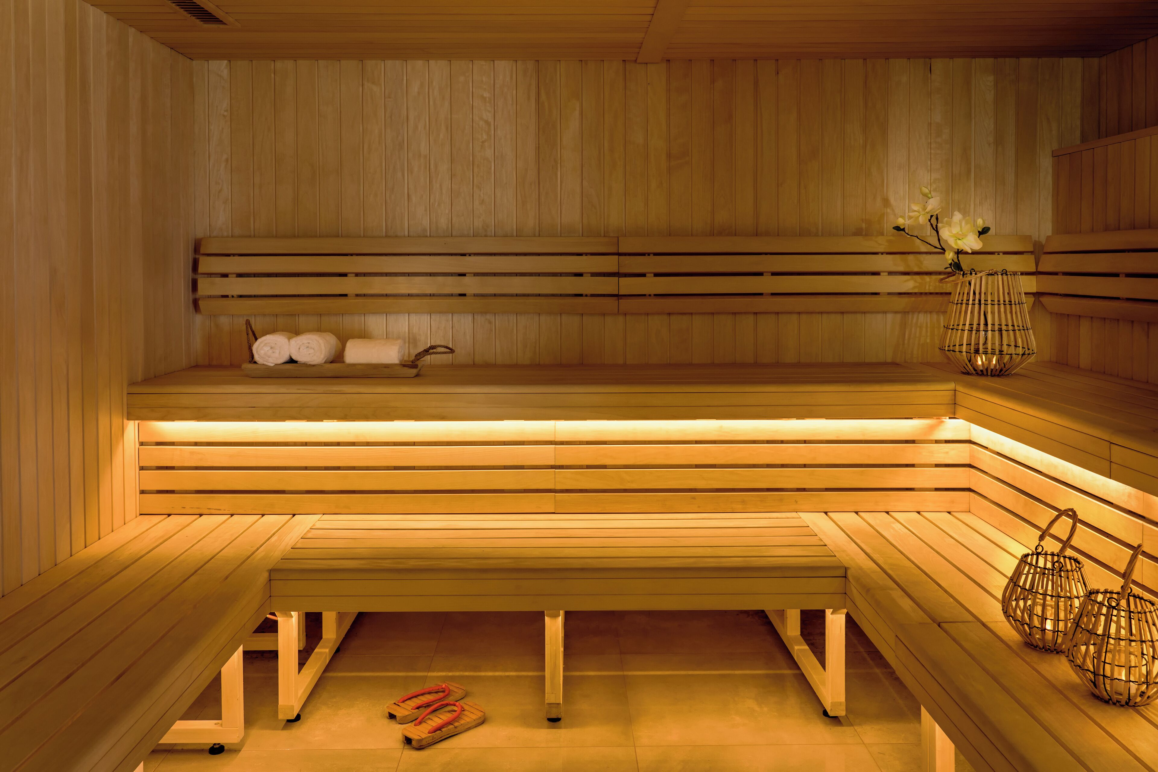 A wooden sauna with benches and a bench seat, a small table with a vase of flowers, and a pair of slippers on the floor.