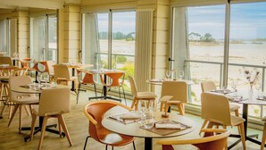 A restaurant with tables and chairs, large windows with a view of the beach.