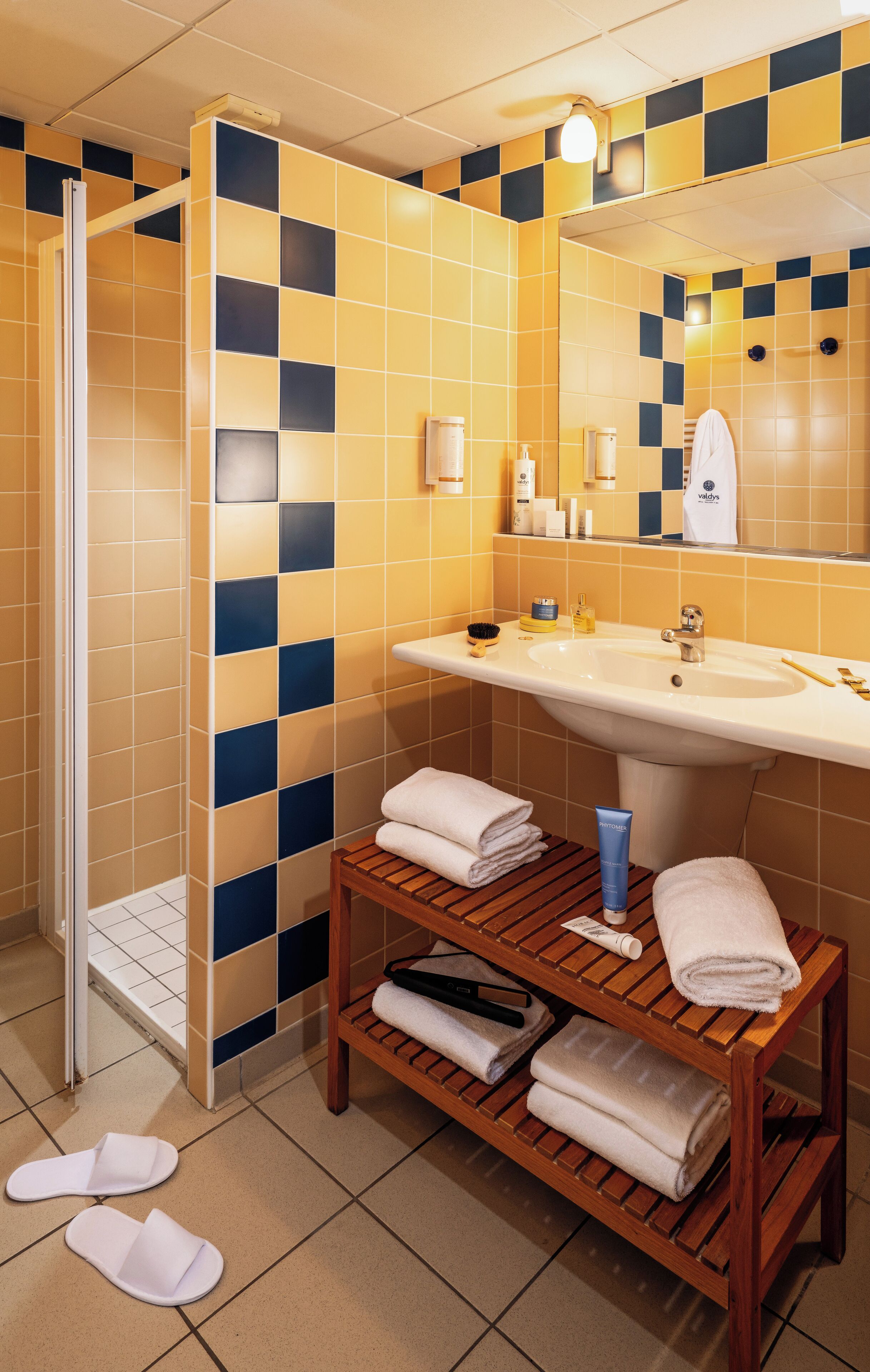 A bathroom with a checkered yellow and blue tile wall, a wooden bench with towels, a sink, and a shower area.