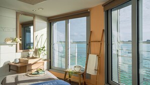 A bedroom with a large window, a bed, a wooden dresser, and a mirror.
