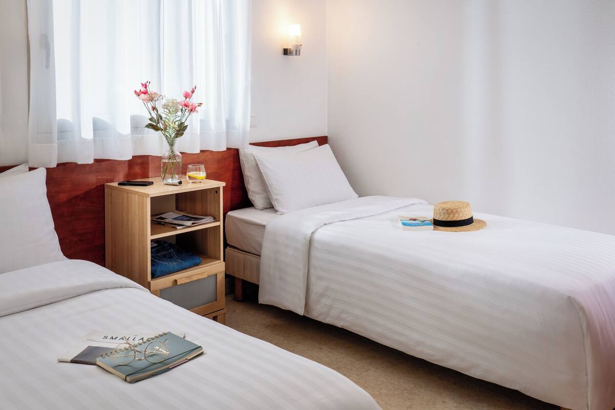 Ein Hotelzimmer mit zwei Betten, einem Nachttisch mit Lampe, einem kleinen Regal mit Büchern und einem Glas sowie einem Fenster mit durchsichtigen Vorhängen.