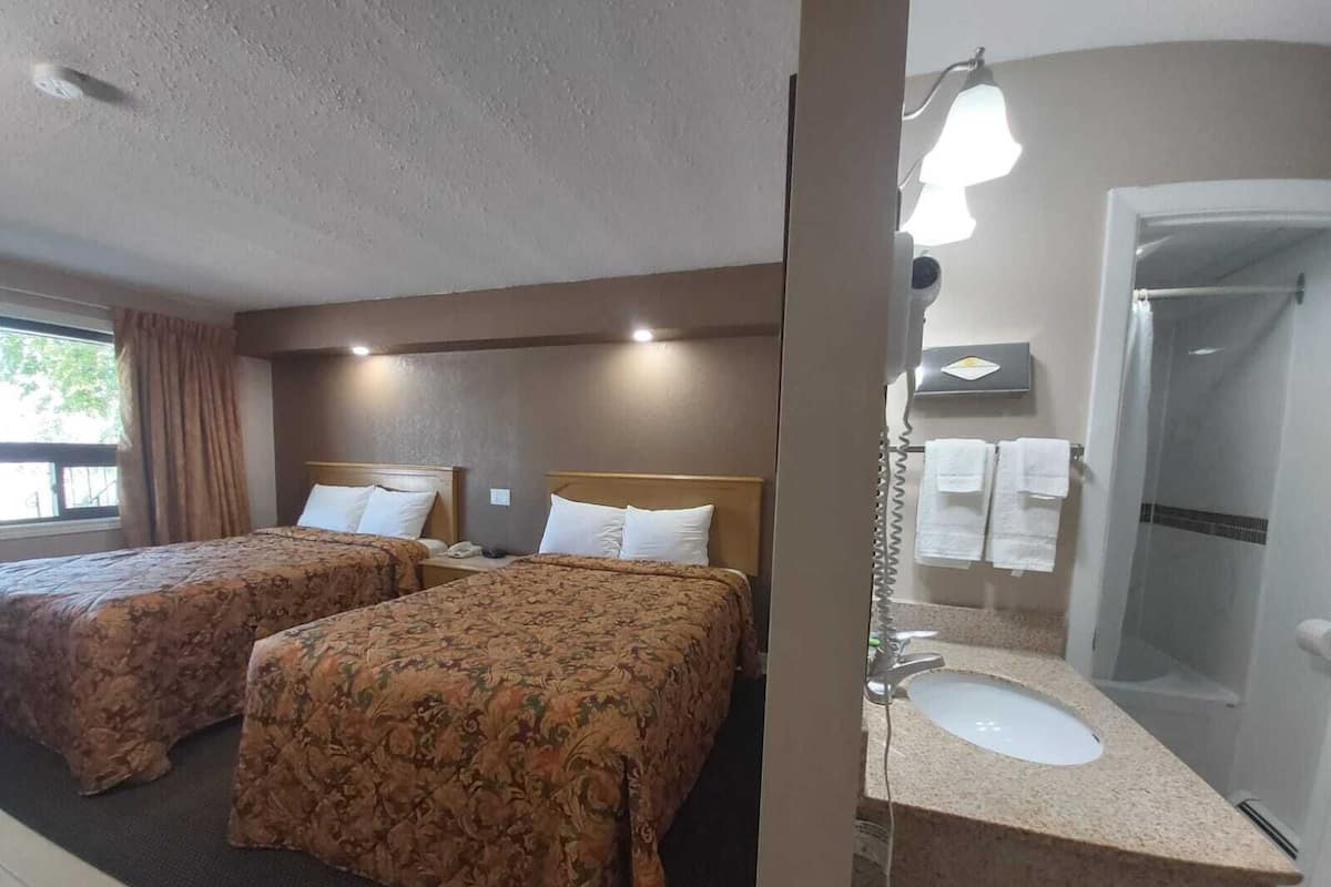 A hotel room with two beds, a bathroom with a sink and mirror, and a shower.