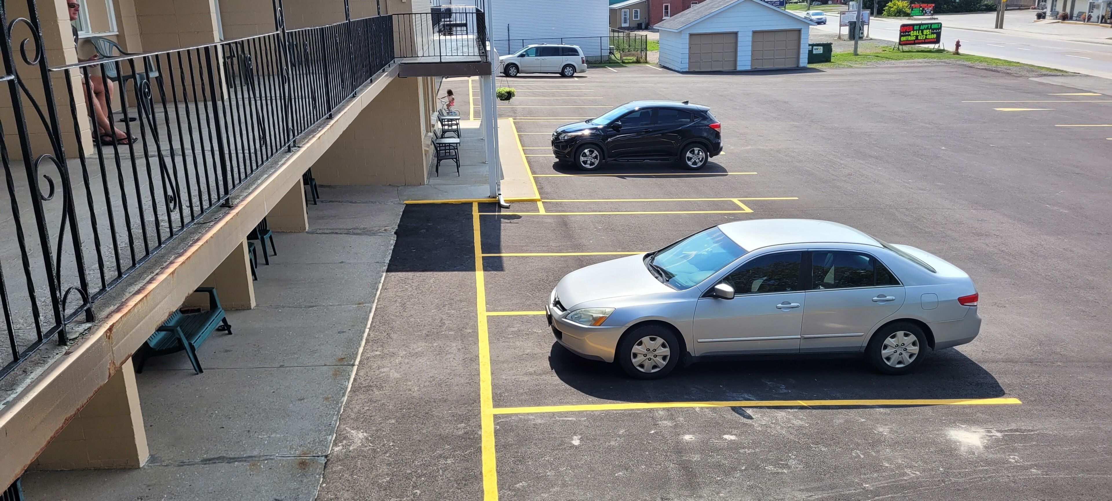 Un parking avec une voiture garée dans un espace délimité, un immeuble avec un balcon et un petit abri de jardin à l’arrière-plan.
