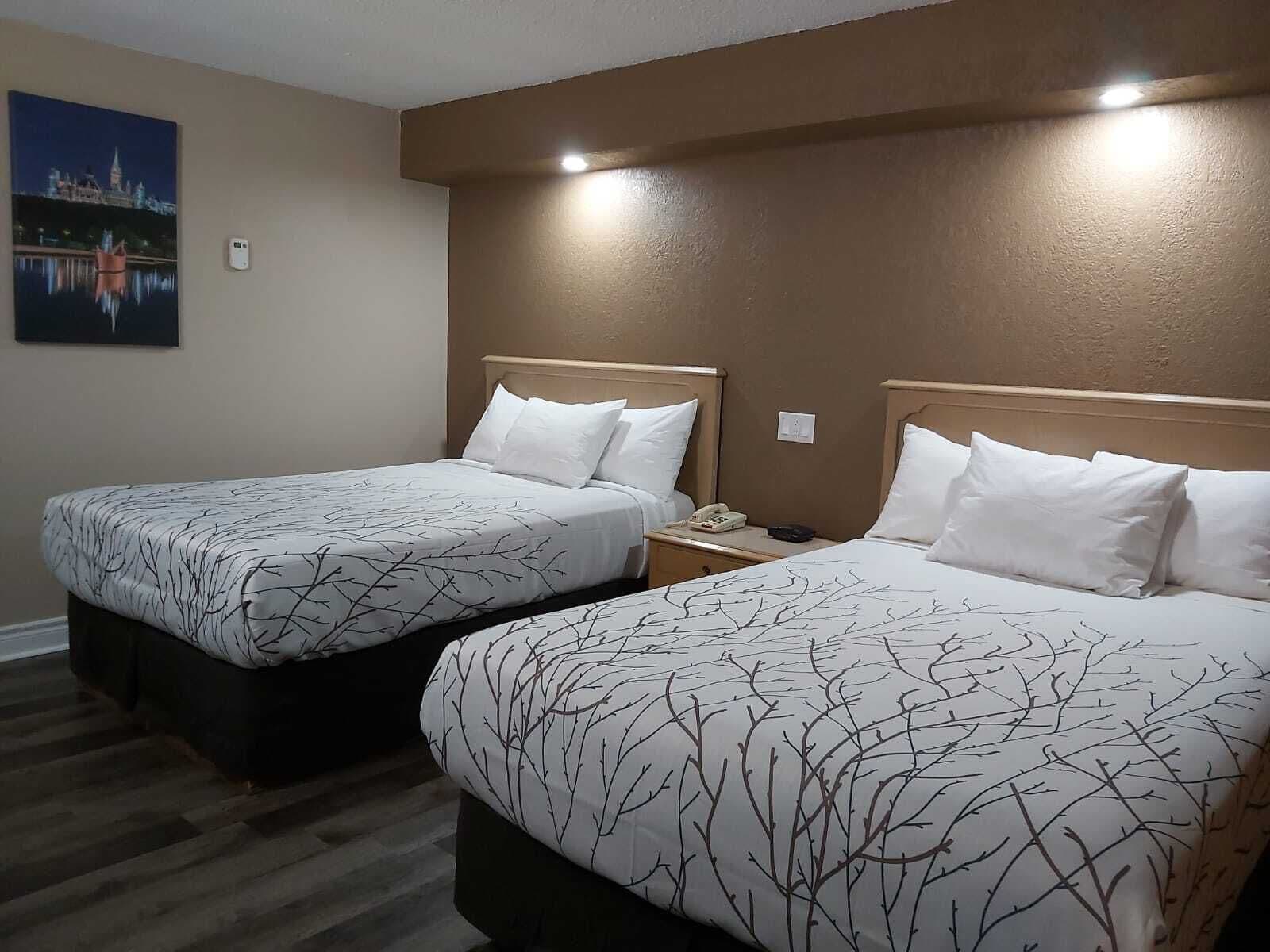 Habitación de hotel con dos camas, cada una con una colcha estampada y almohadas blancas. Hay una mesita de noche con un teléfono y una pequeña mesa entre las camas. Un cuadro de un paisaje urbano adorna la pared.
