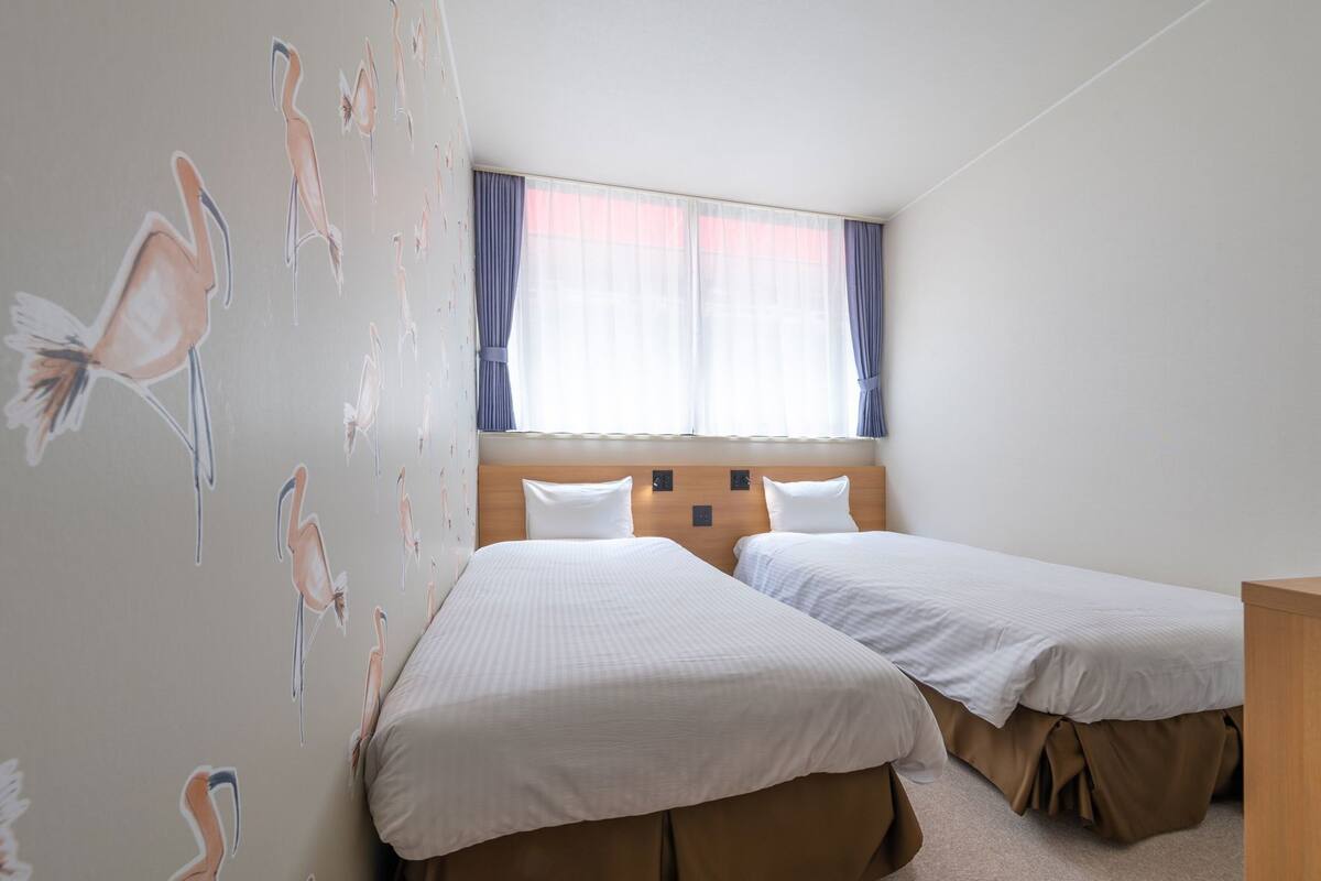A hotel room with two beds, a wooden headboard, a window with curtains, and wall art of flamingos.