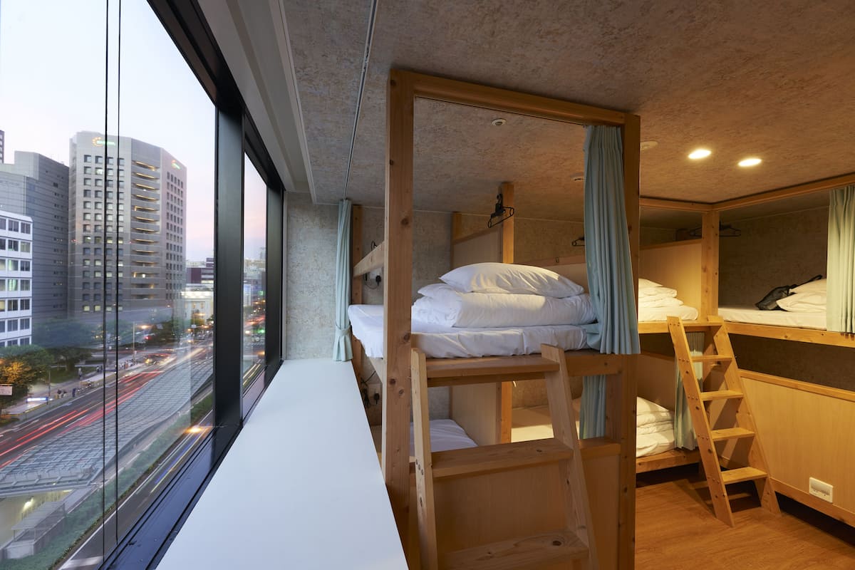 A compact hotel room with a bunk bed, a wooden ladder, and a window overlooking a cityscape.
