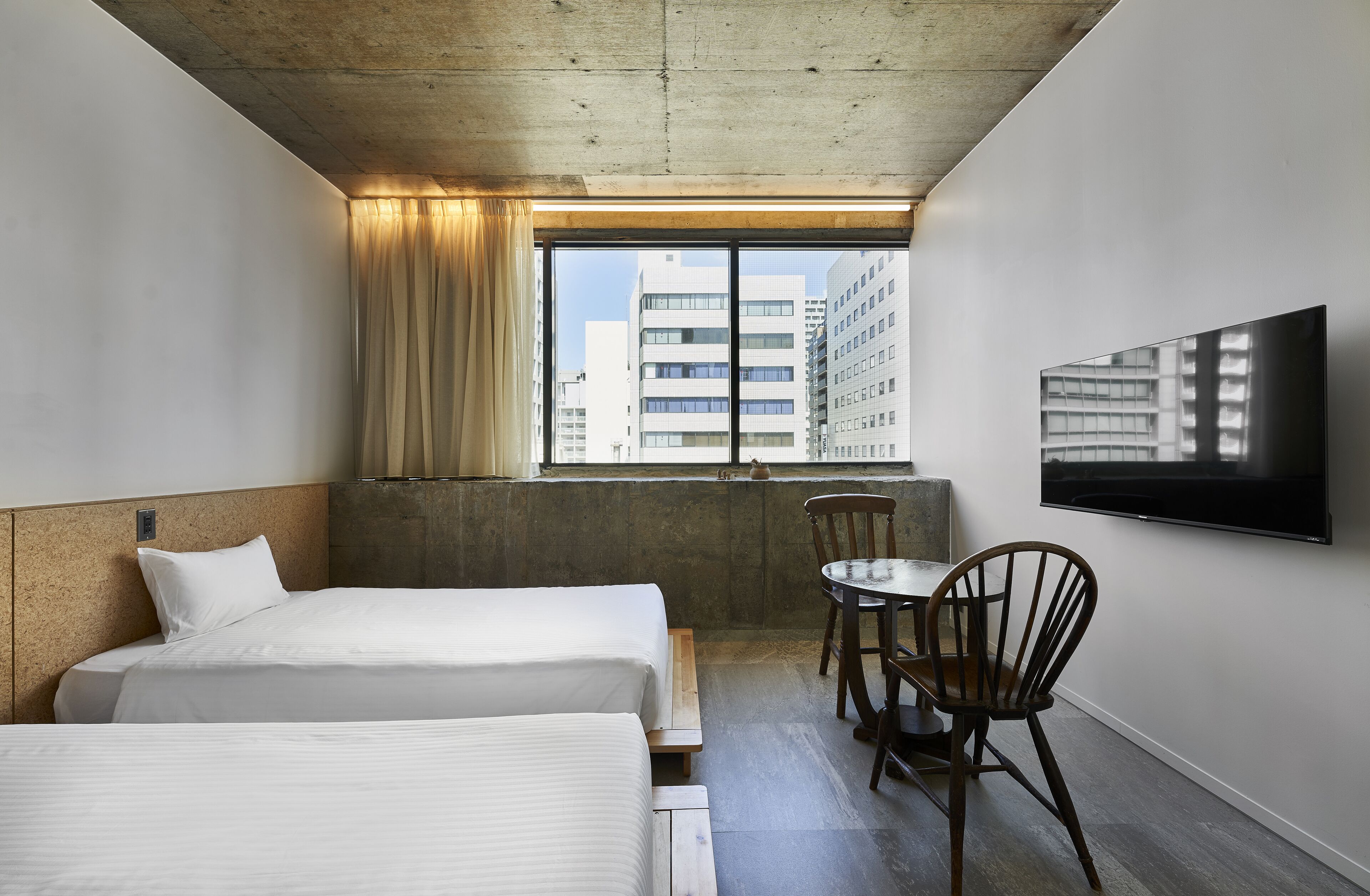 A hotel room with two beds, a TV, a window with a view of buildings, and a small table with a chair.
