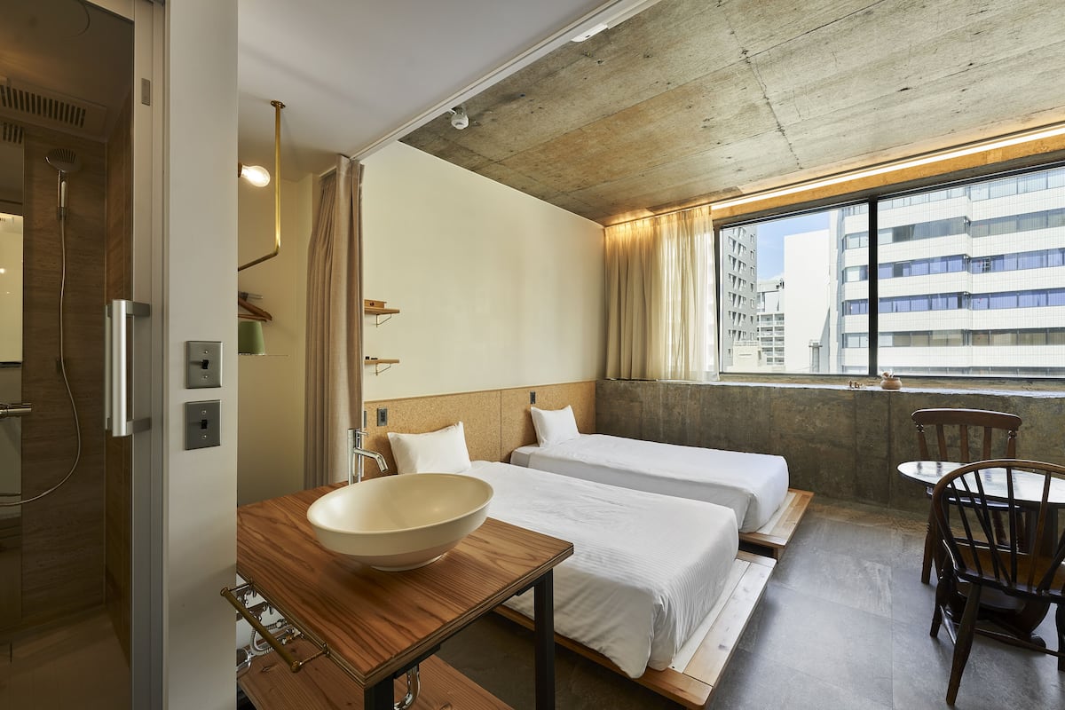 A hotel room with a wooden table, a bowl sink, two beds, a chair, and a view of the city through large windows.