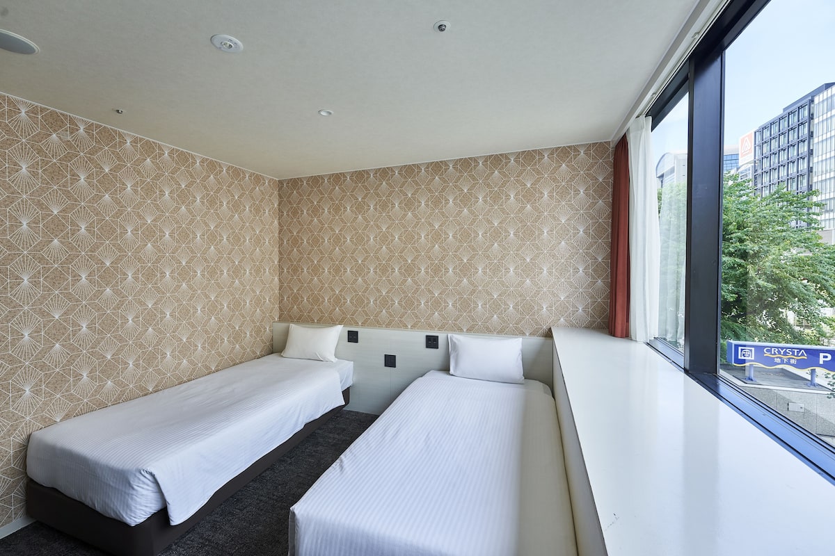 A hotel room with two single beds, a large window with a view of buildings, and a patterned wall.