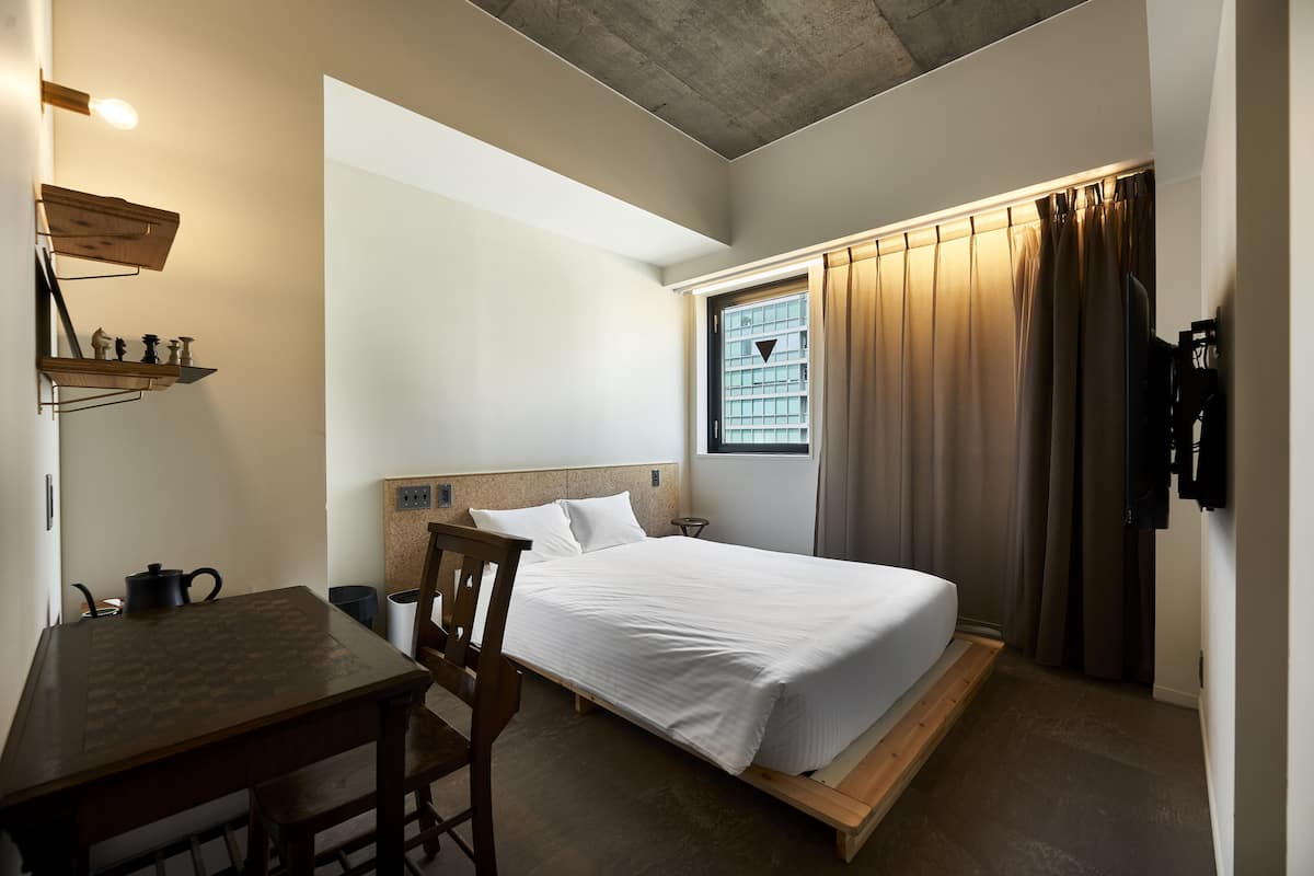 A compact bedroom with a bed, a small table, and a chair. There is a window with curtains and a shelf with decorative items.