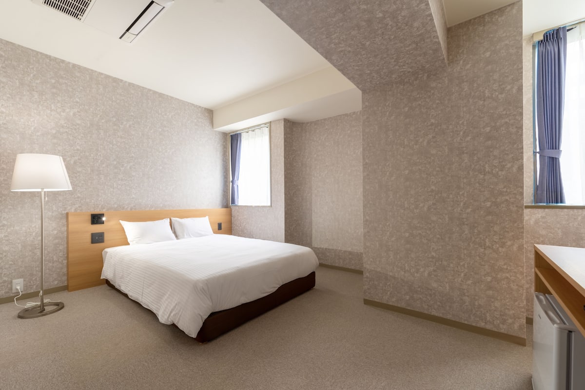 A modern hotel room with a large bed, a bedside lamp, a small desk, and a window with curtains.