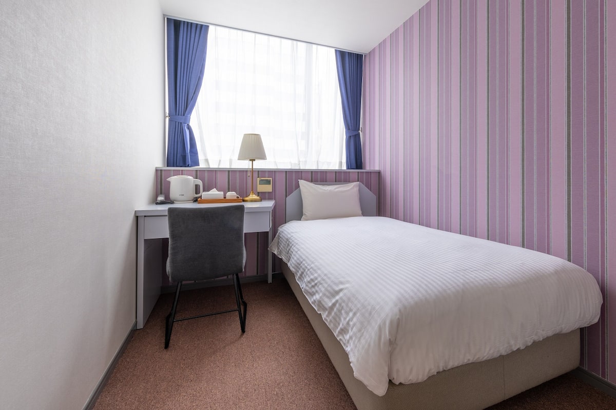 A hotel room with a bed, a desk, a chair, a lamp, and a window with curtains.