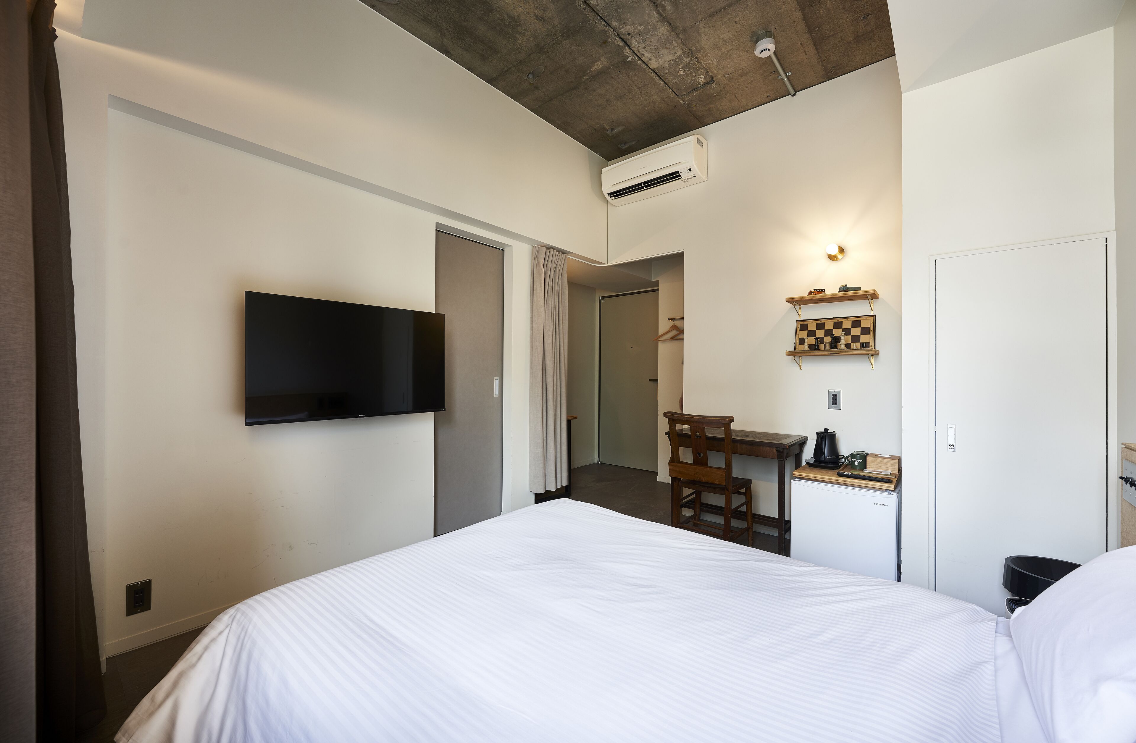 A hotel room with a bed, a desk with a chair, a television mounted on the wall, and a small refrigerator.