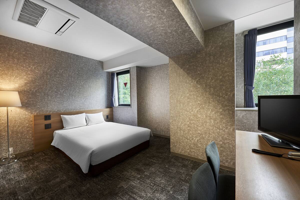 A hotel room with a bed, a desk with a TV, a lamp, and a window with curtains.