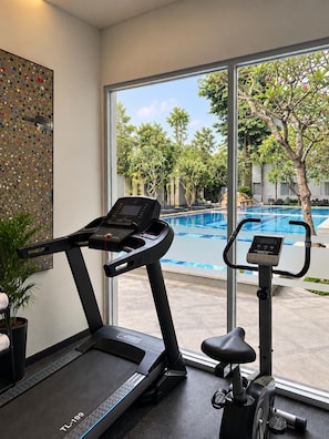 A gym with a treadmill and an elliptical machine, a large window with a view of a swimming pool and trees, and a decorative wall piece. - Gets Premiere Yogyakarta (Depok)