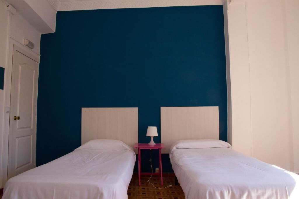 Una habitación con dos camas individuales, una mesita de noche con lámpara y una puerta.