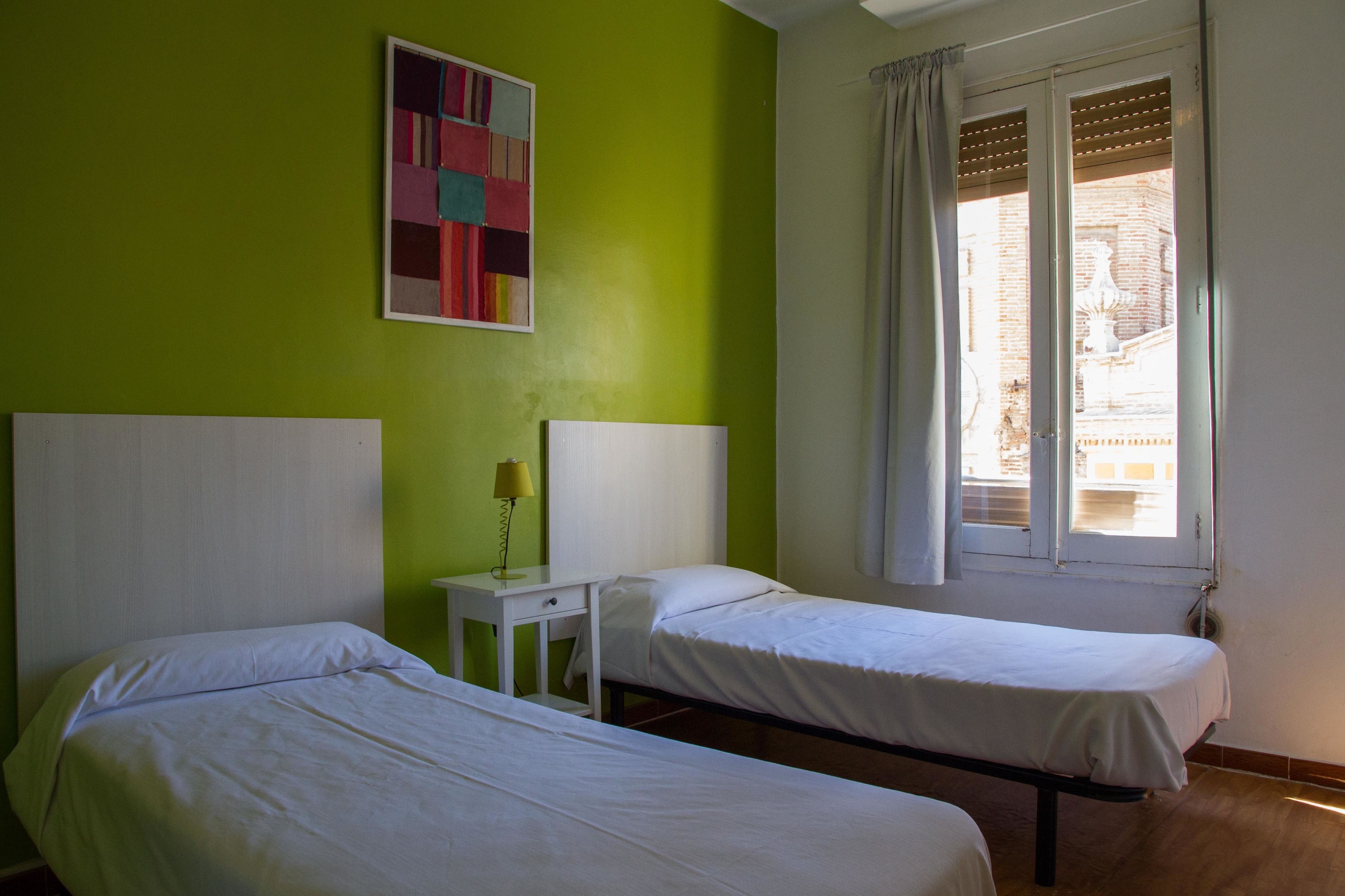 A room with two beds, a small table with a lamp, and a window with curtains.