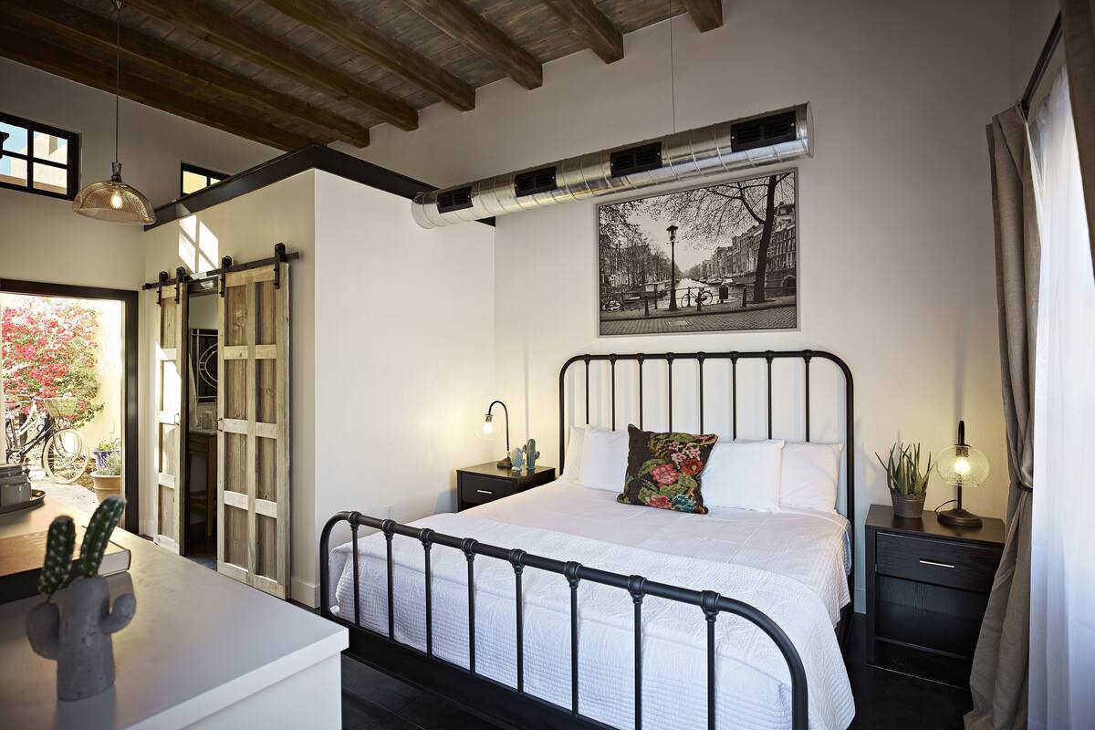 A bedroom with a bed, bedside tables, a nightstand, a potted plant, a hanging light fixture, and a window with a view of a street and buildings.