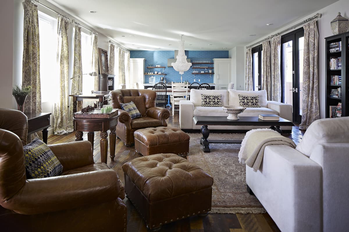 A spacious living room with leather armchairs, a white sofa, and a coffee table. There is a dining area with a chandelier and a kitchen in the background.