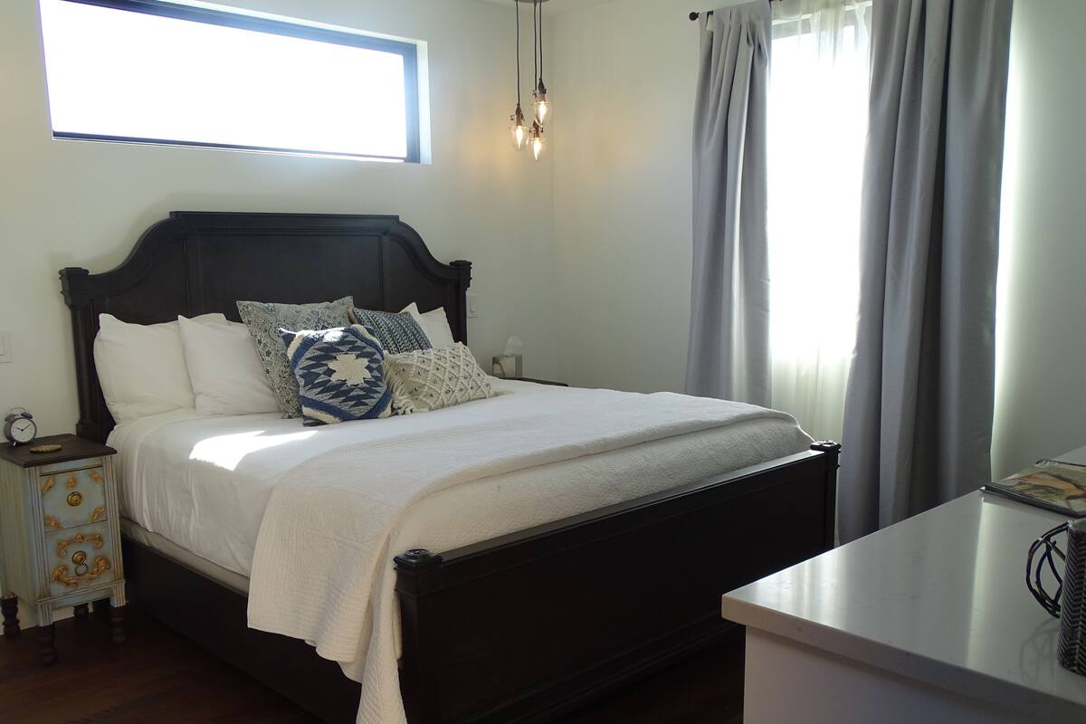 A neatly made bed with a dark headboard, white bedding, and decorative pillows. A small bedside table with a clock and a decorative item is visible.