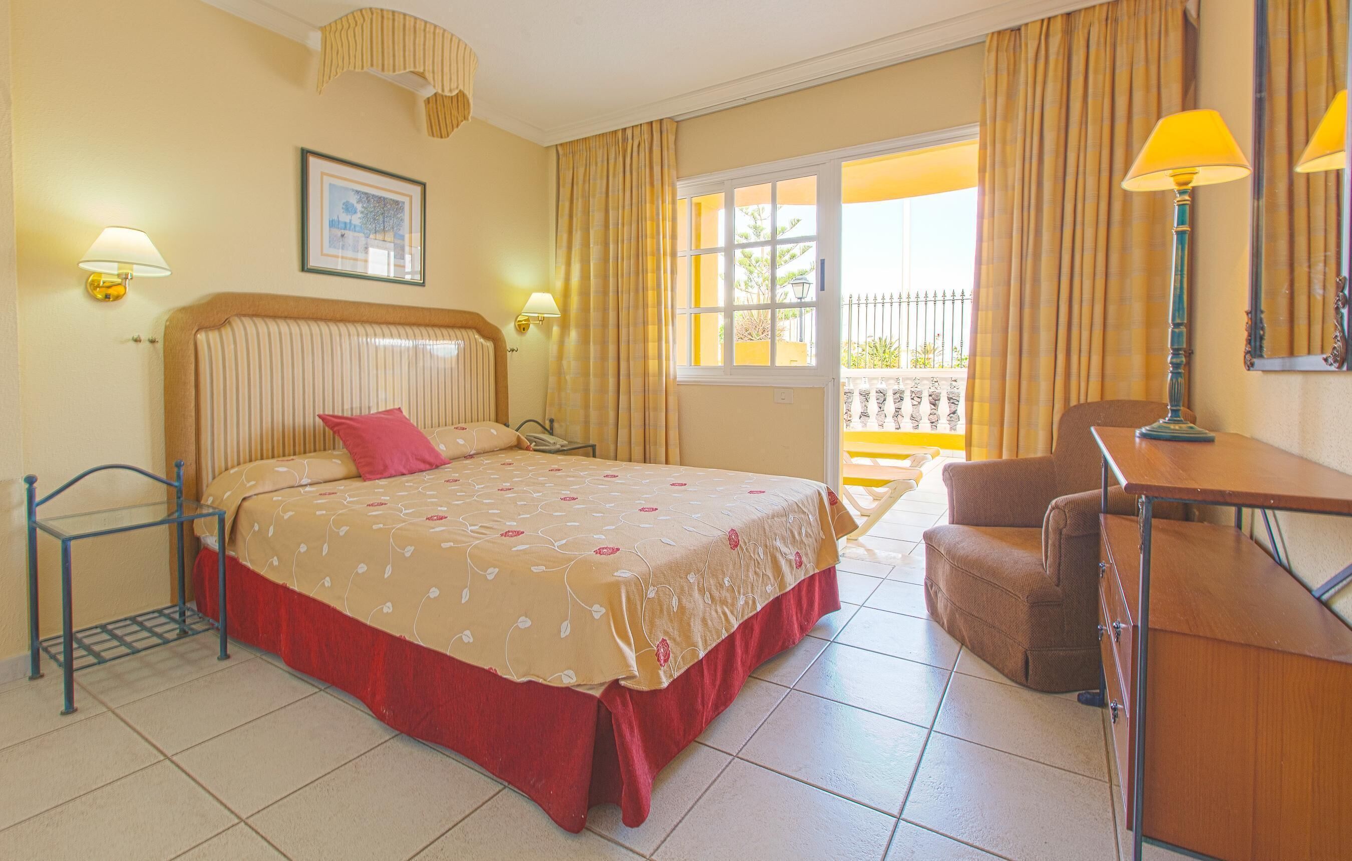 A hotel room with a bed, a desk, a chair, a window with curtains, a lamp, and a picture on the wall.