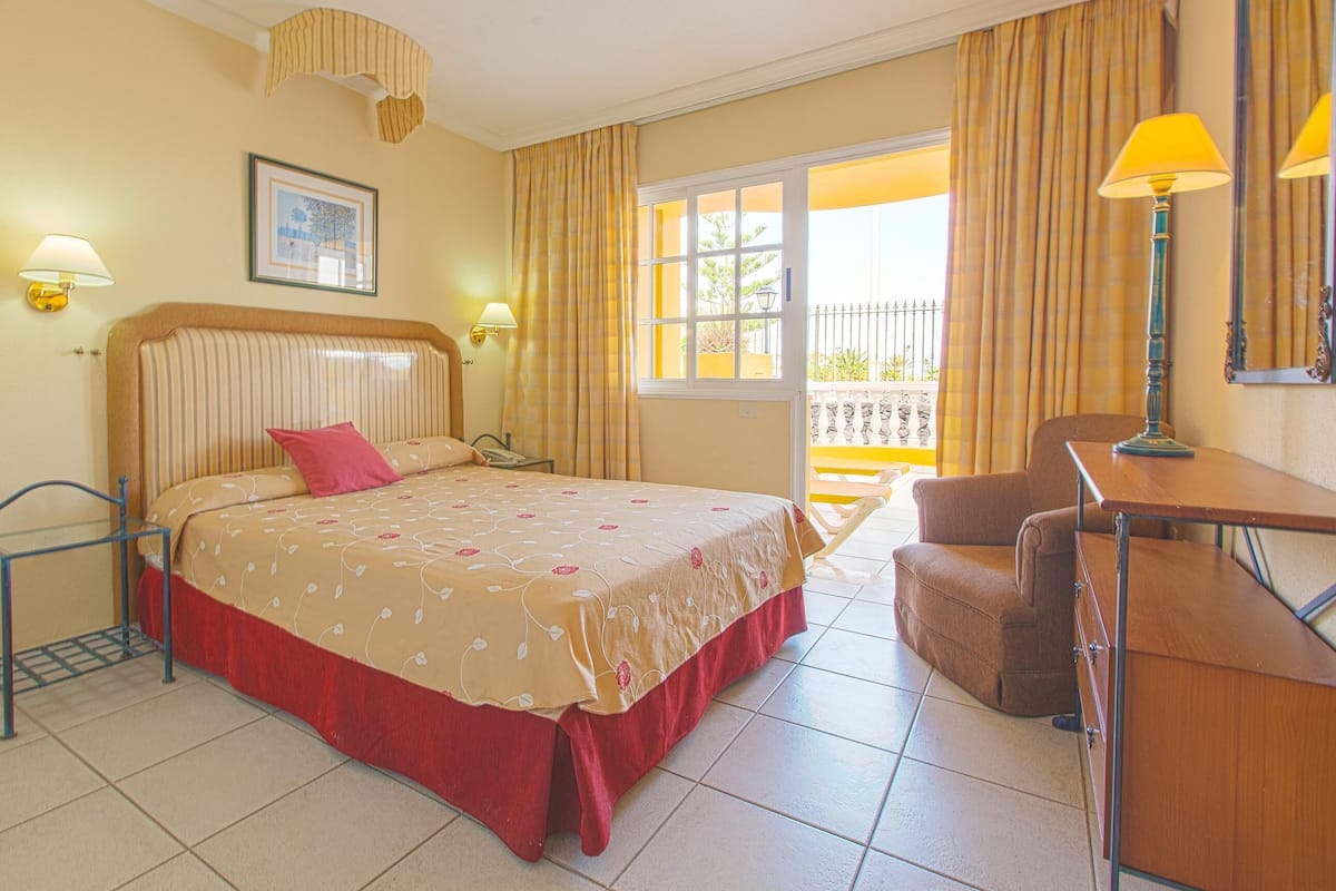 A hotel room with a bed, a desk, a chair, a window with curtains, a lamp, and a picture on the wall.