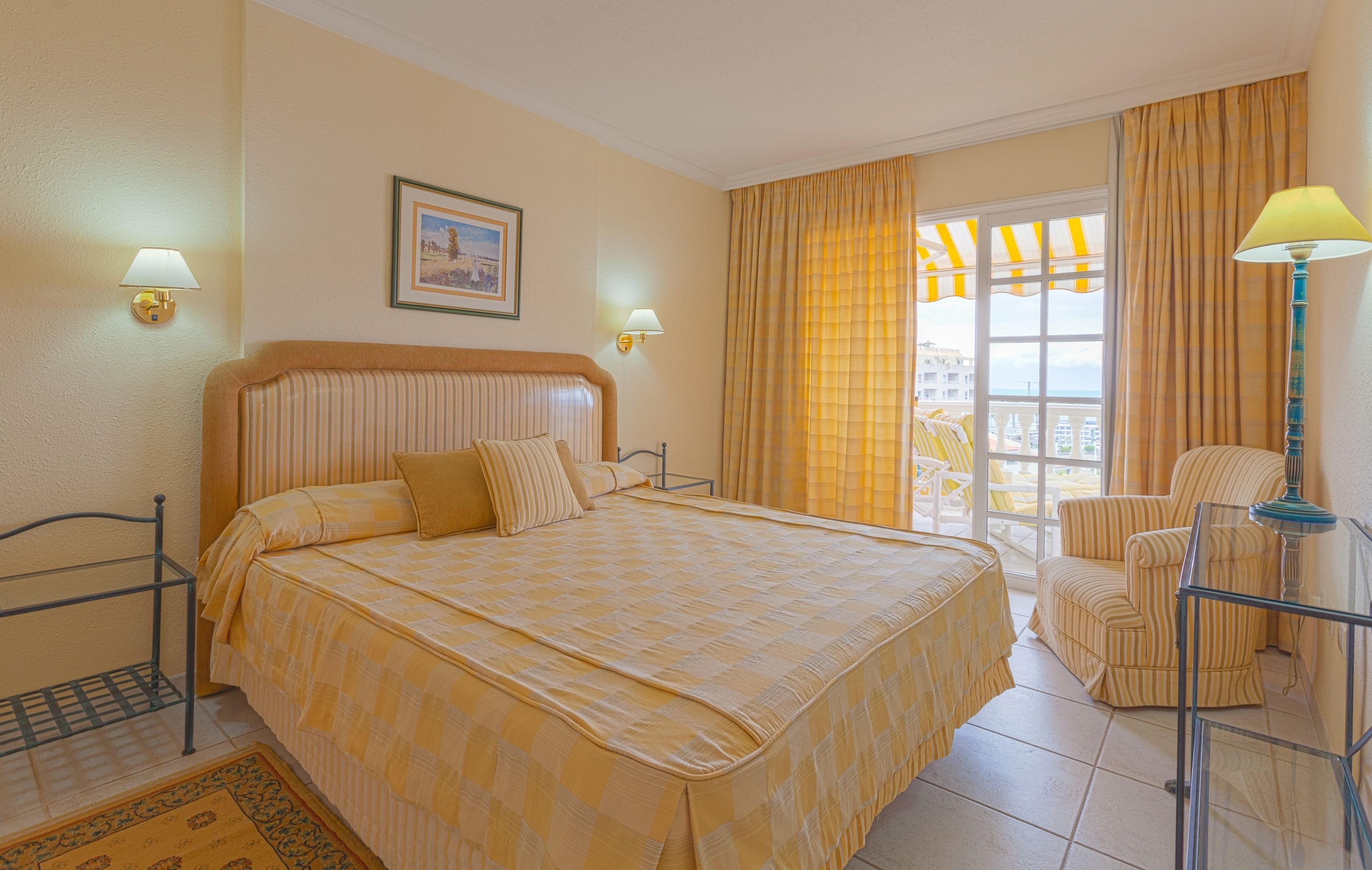 Ein Hotelzimmer mit einem großen Bett, zwei Nachttischlampen, einem Sessel, einem Balkon mit gelben Stühlen und Meerblick.