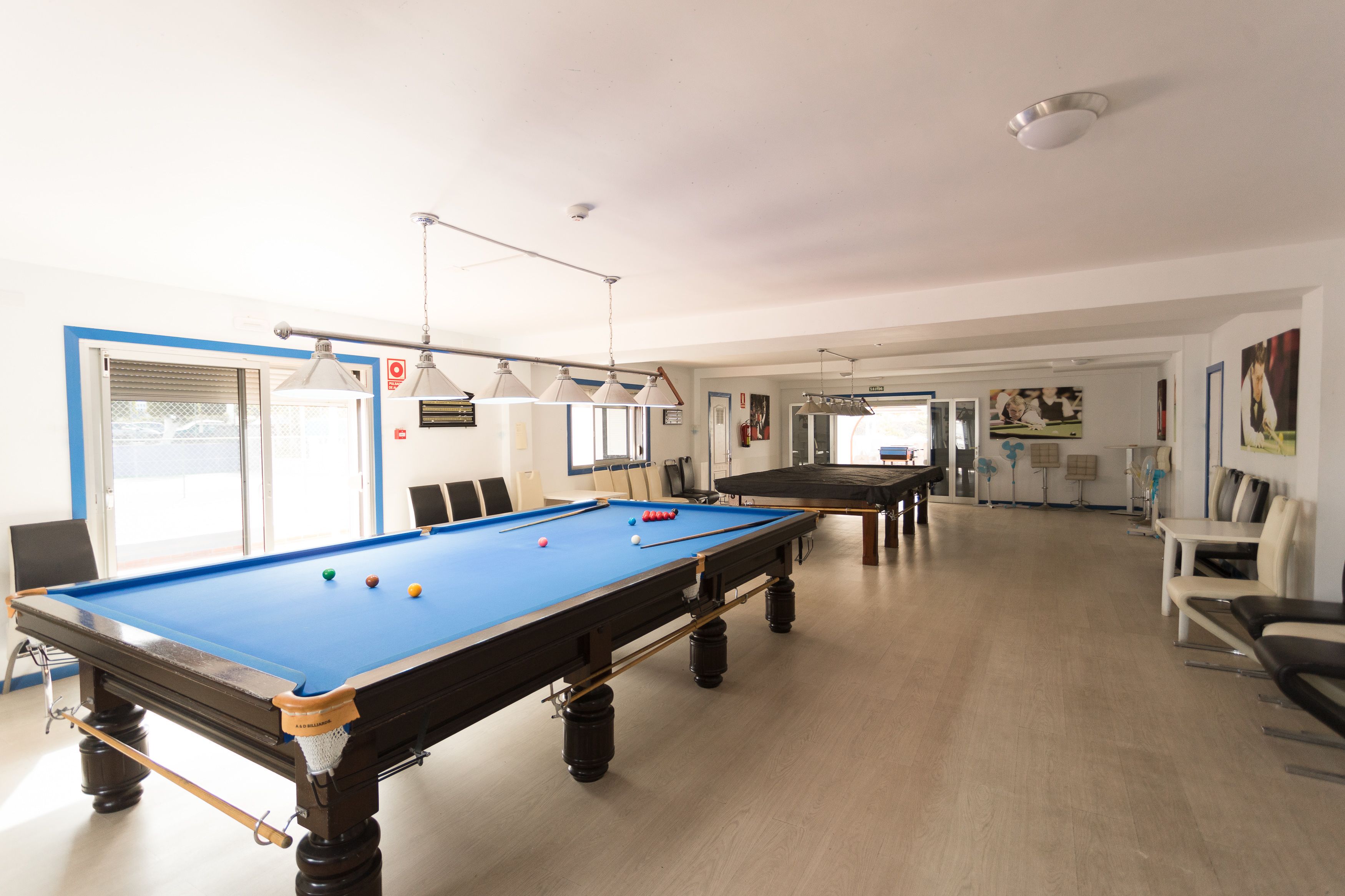 Une table de billard dans une pièce bien éclairée, avec un sol carrelé de carreaux bleus, un coin salon avec des chaises et une porte dotée d’une fenêtre.