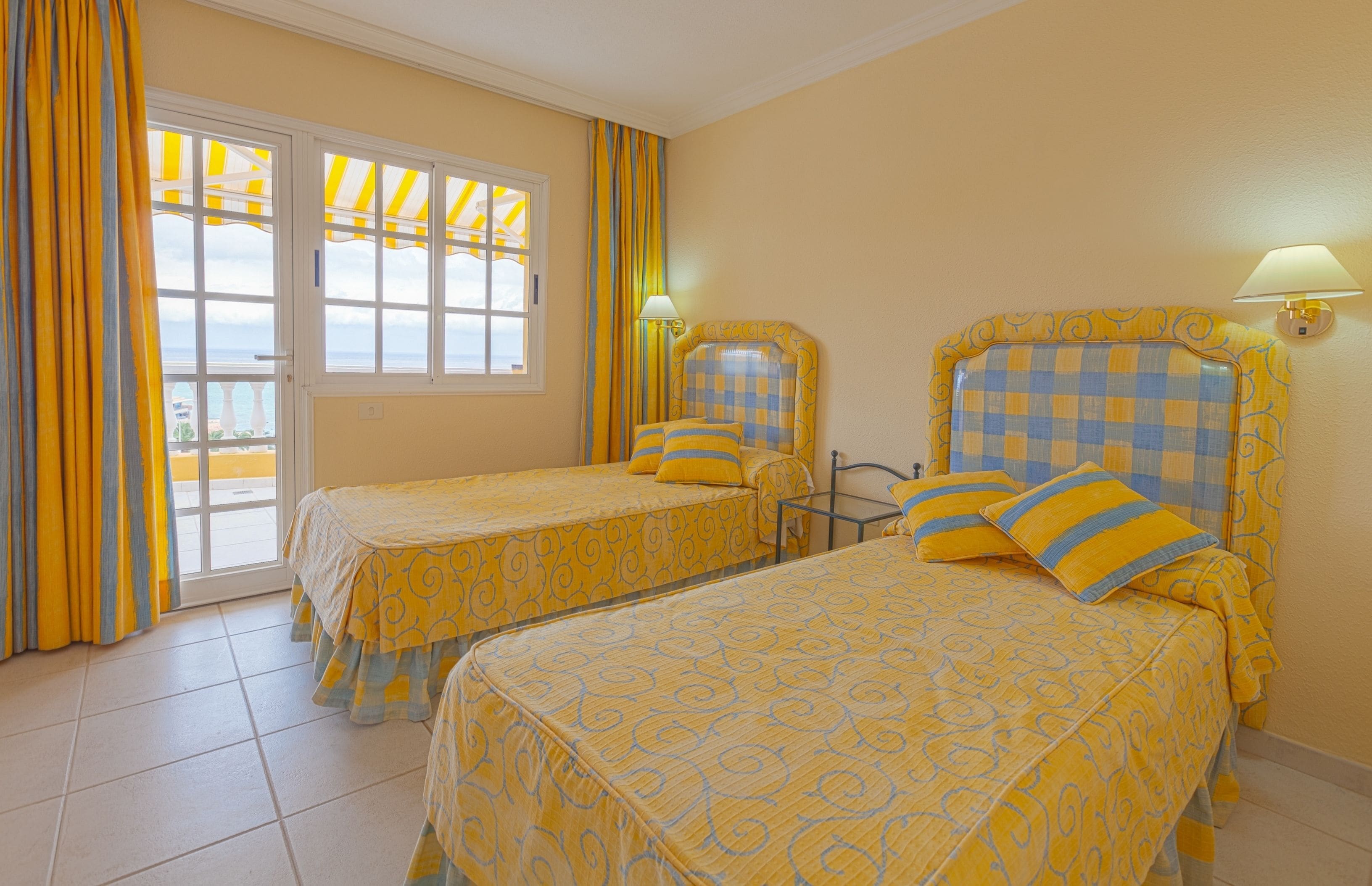 Une chambre d’hôtel avec deux lits, un balcon avec des rideaux jaunes et une vue sur la mer.