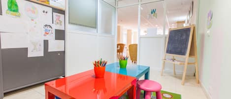 Un aula con una mesa roja, sillas azules y rosadas, una pizarra y dibujos infantiles en la pared.