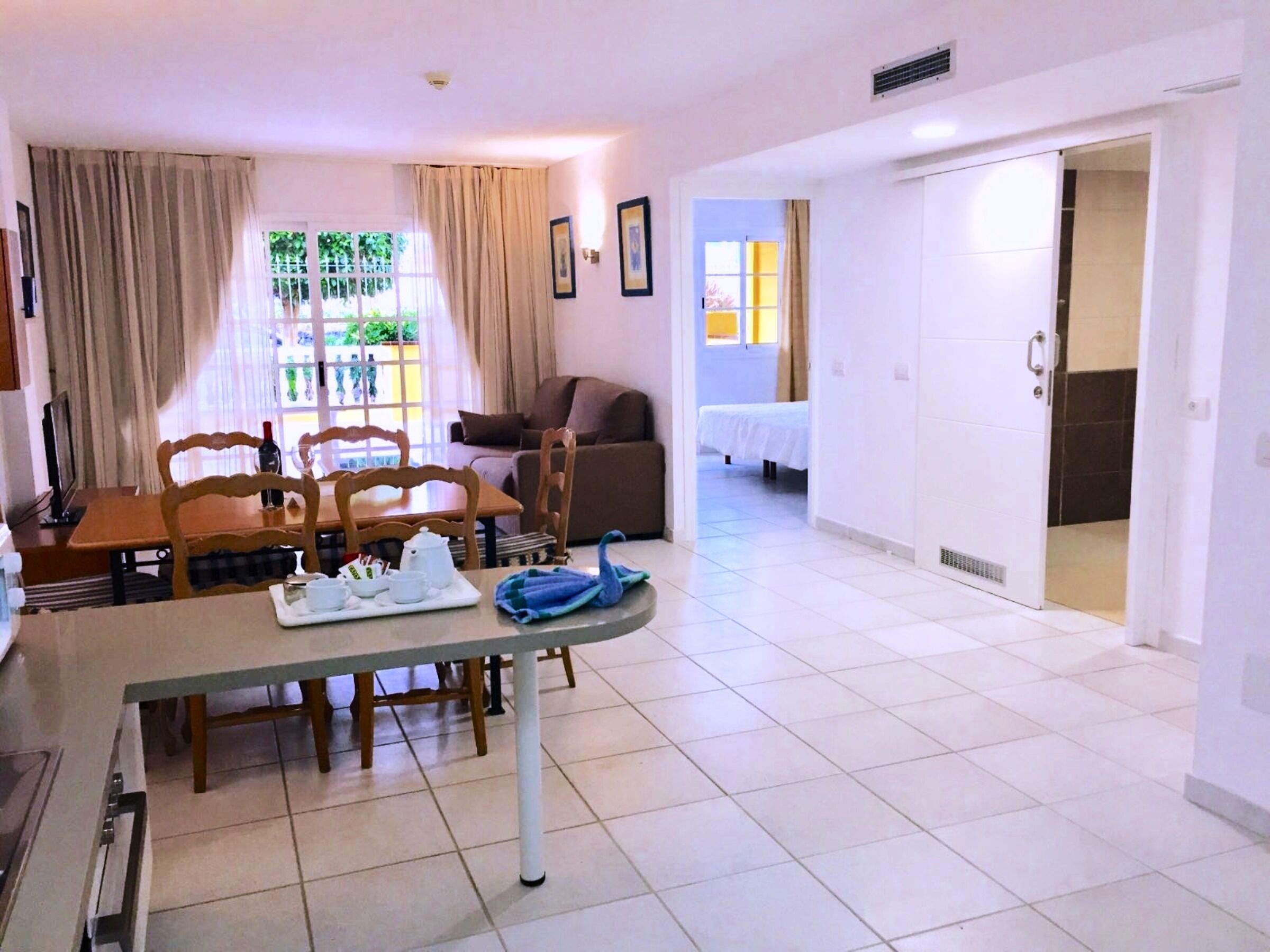 A dining area with a table, chairs, and a window with curtains. There is a bed in the background and a door leading to another room.