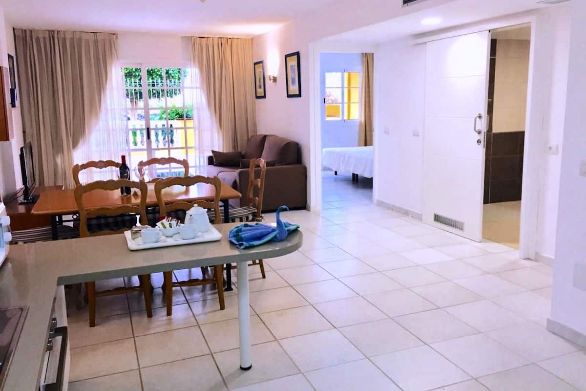 A dining area with a table, chairs, and a window with curtains. There is a bed in the background and a door leading to another room.