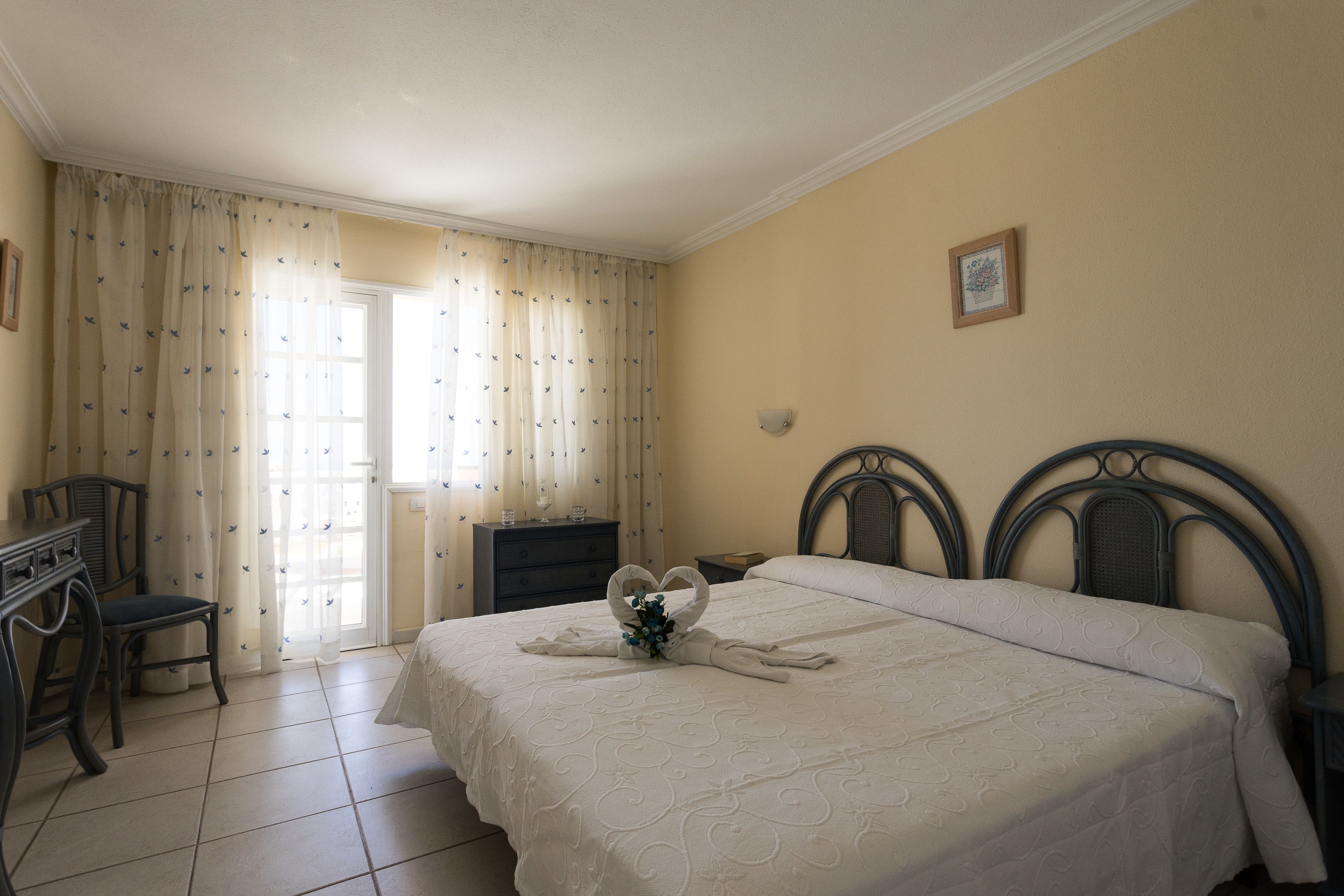 A neatly made bed with a heart-shaped towel and flowers, a desk with a chair, and a door with curtains.