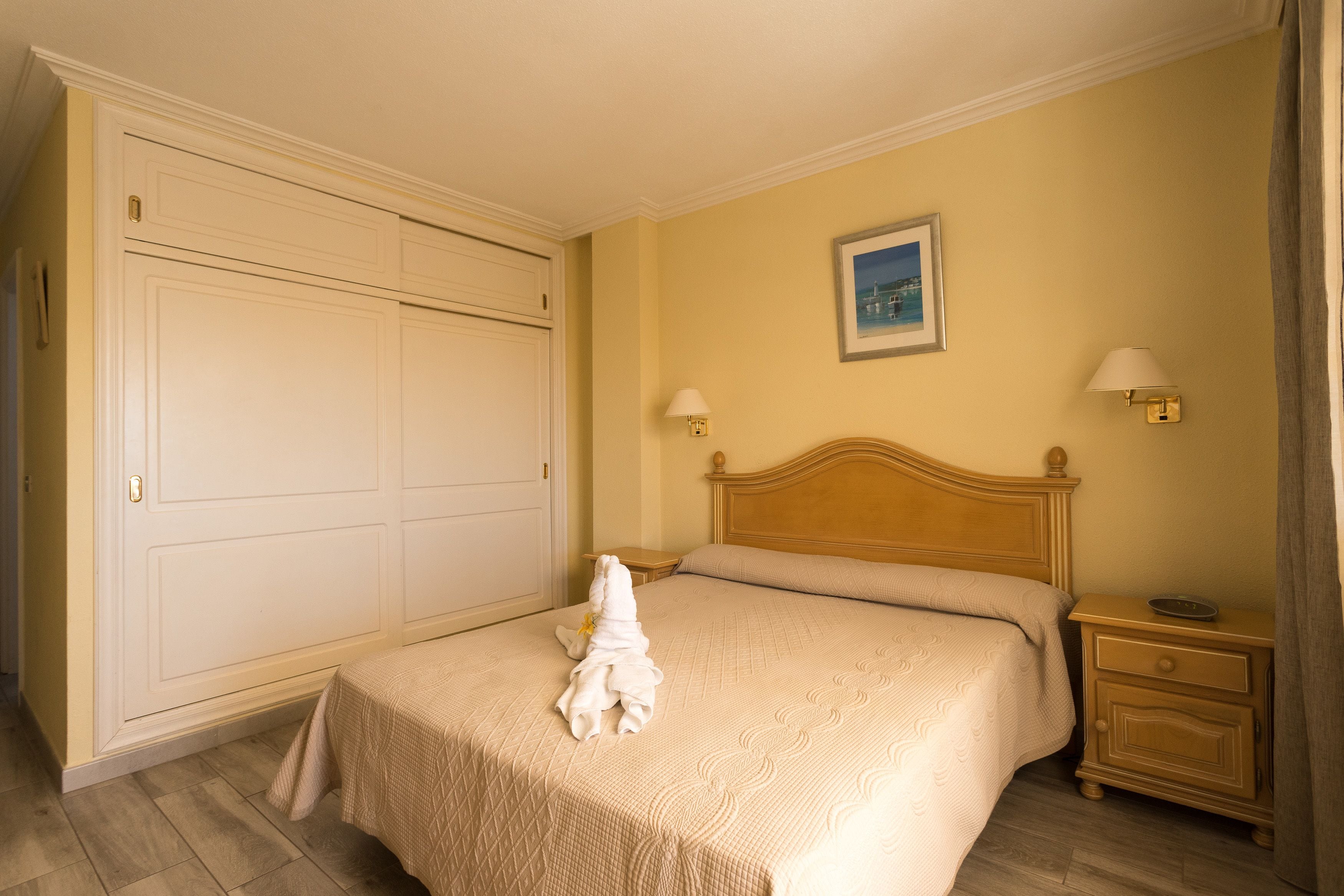 A hotel room with a large wardrobe, a bed with a neatly folded white towel, two bedside lamps, and a framed picture on the wall.