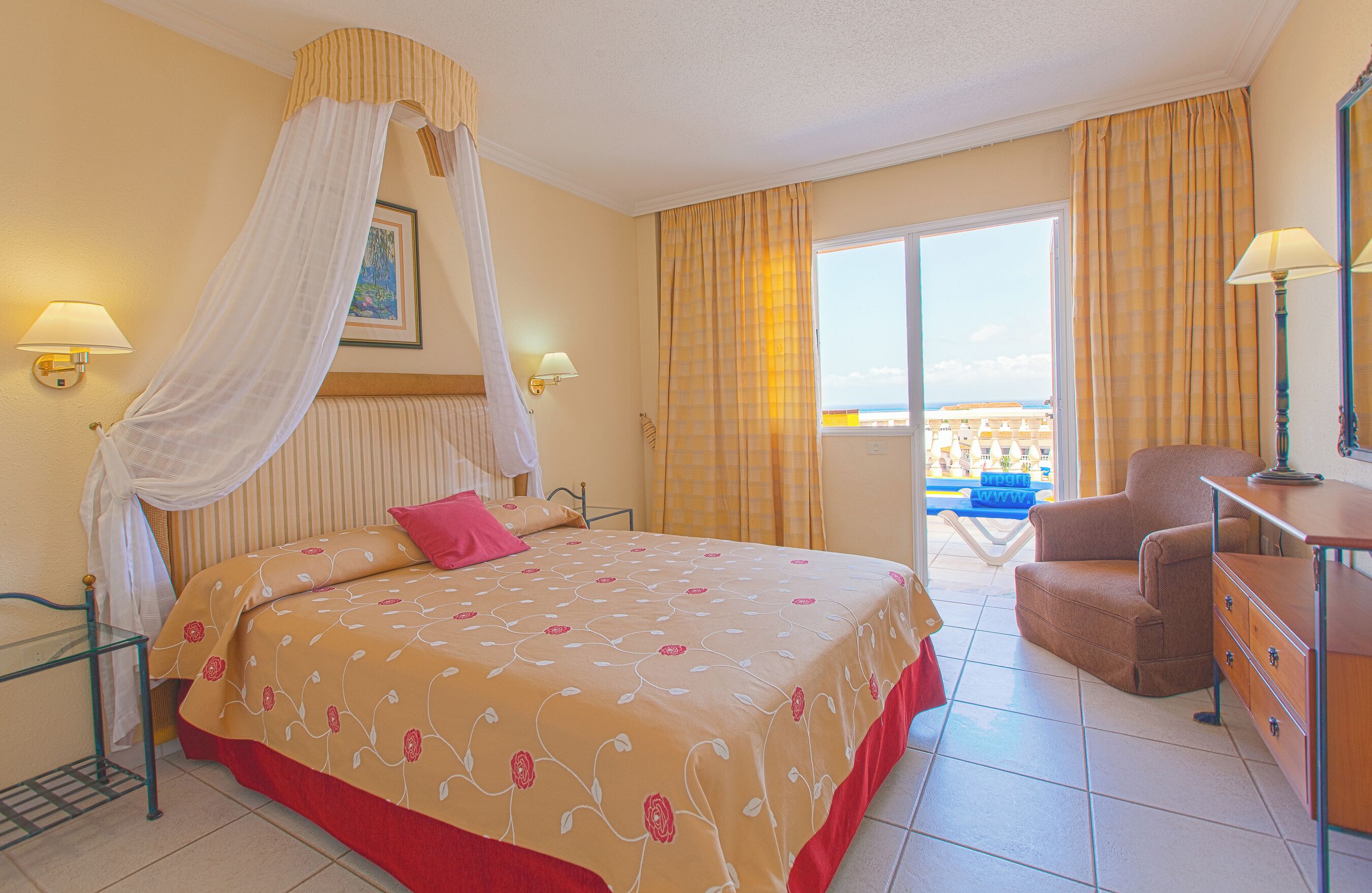 A hotel room with a bed, a chair, a desk, a lamp, and a view of the sea.