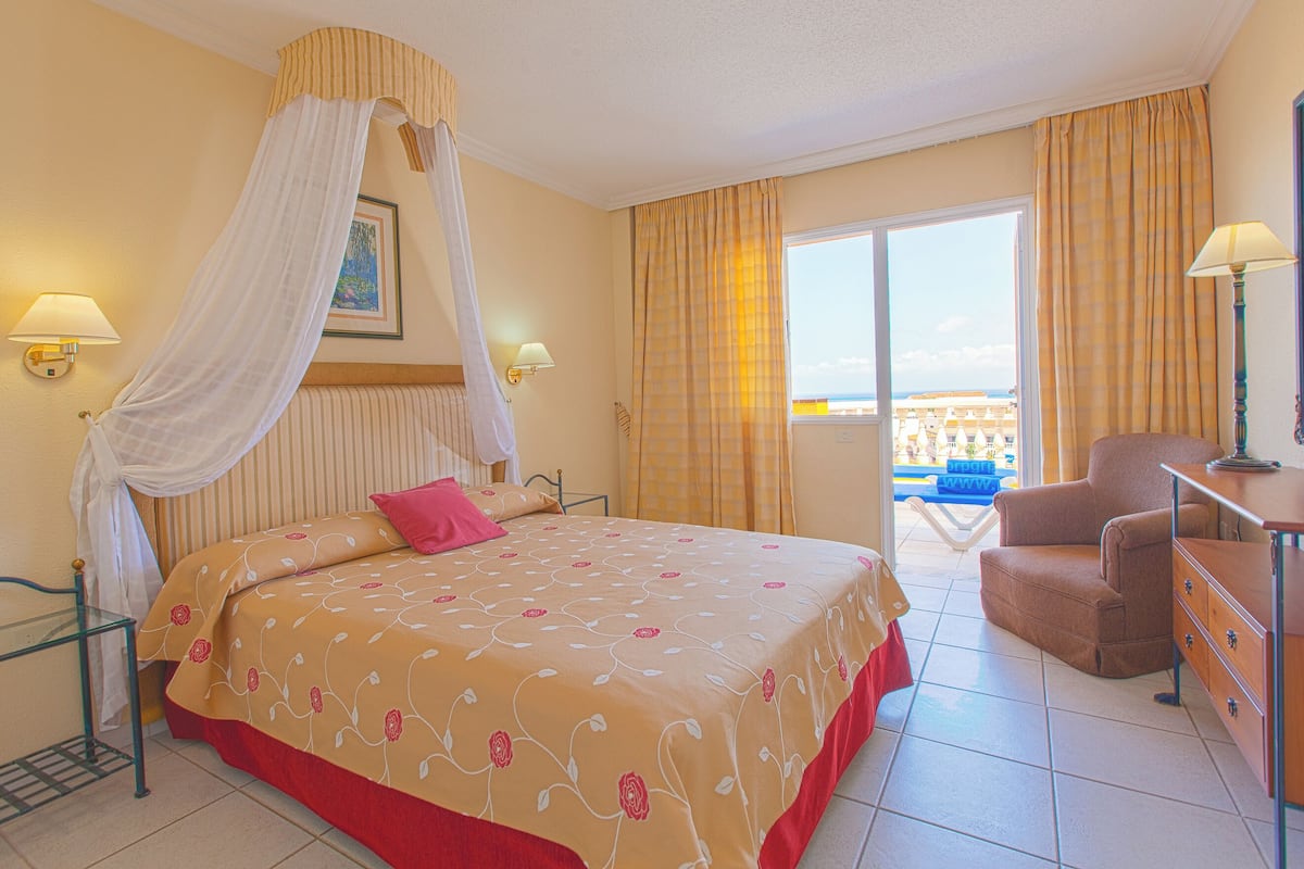 A hotel room with a bed, a chair, a desk, and a balcony with a view.
