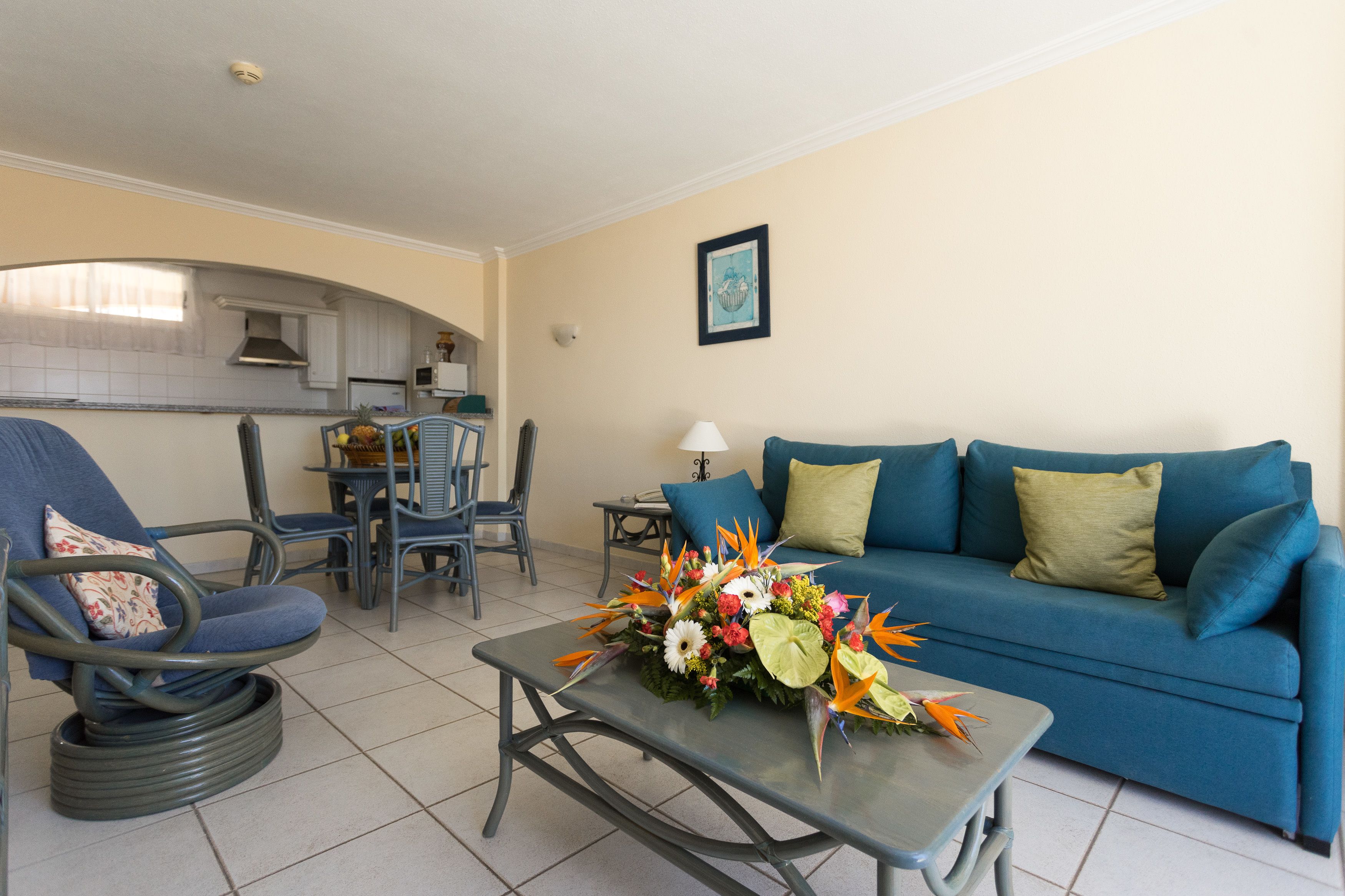 Una sala de estar con un sofá azul, una silla, una mesa de centro con un arreglo floral, y una cocina al fondo.