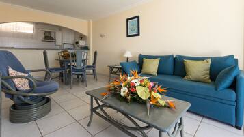 Una sala de estar con un sofá azul, una silla, una mesa de centro con un arreglo floral, y una cocina al fondo.