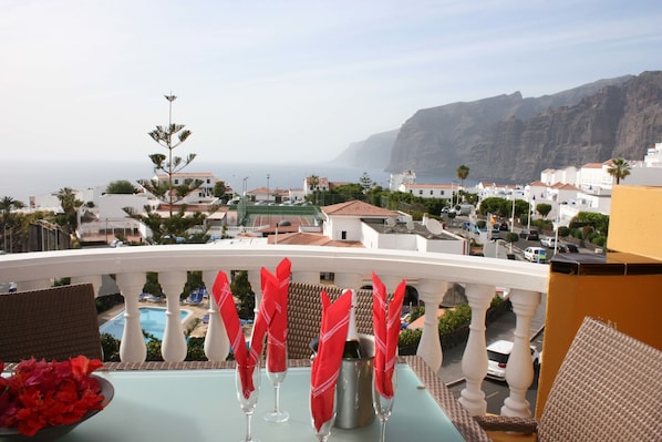A balcony with a view of the sea and buildings, a pool, and red napkins on a table.