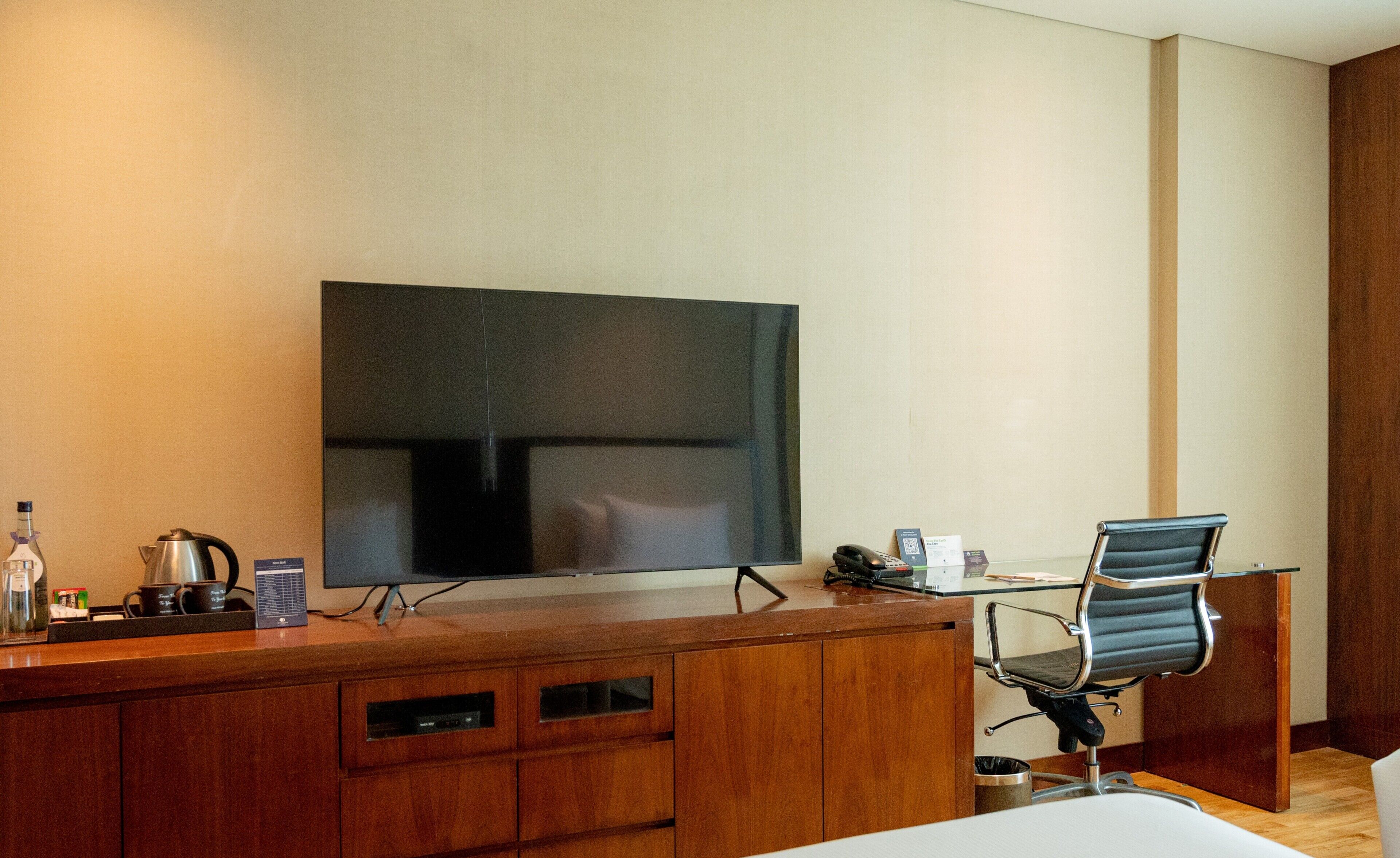 A hotel room with a wooden desk, a flat-screen TV, a chair, a coffee maker, and a trash can.