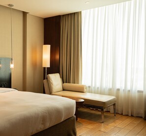 Una habitación de hotel con cama, sofá, lámpara y un amplio ventanal con cortinas.