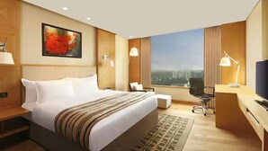 A hotel room with a large bed, a desk with a TV, a chair, and a view of buildings. - Doubletree By Hilton Pune - Chinchwad (Pune)
