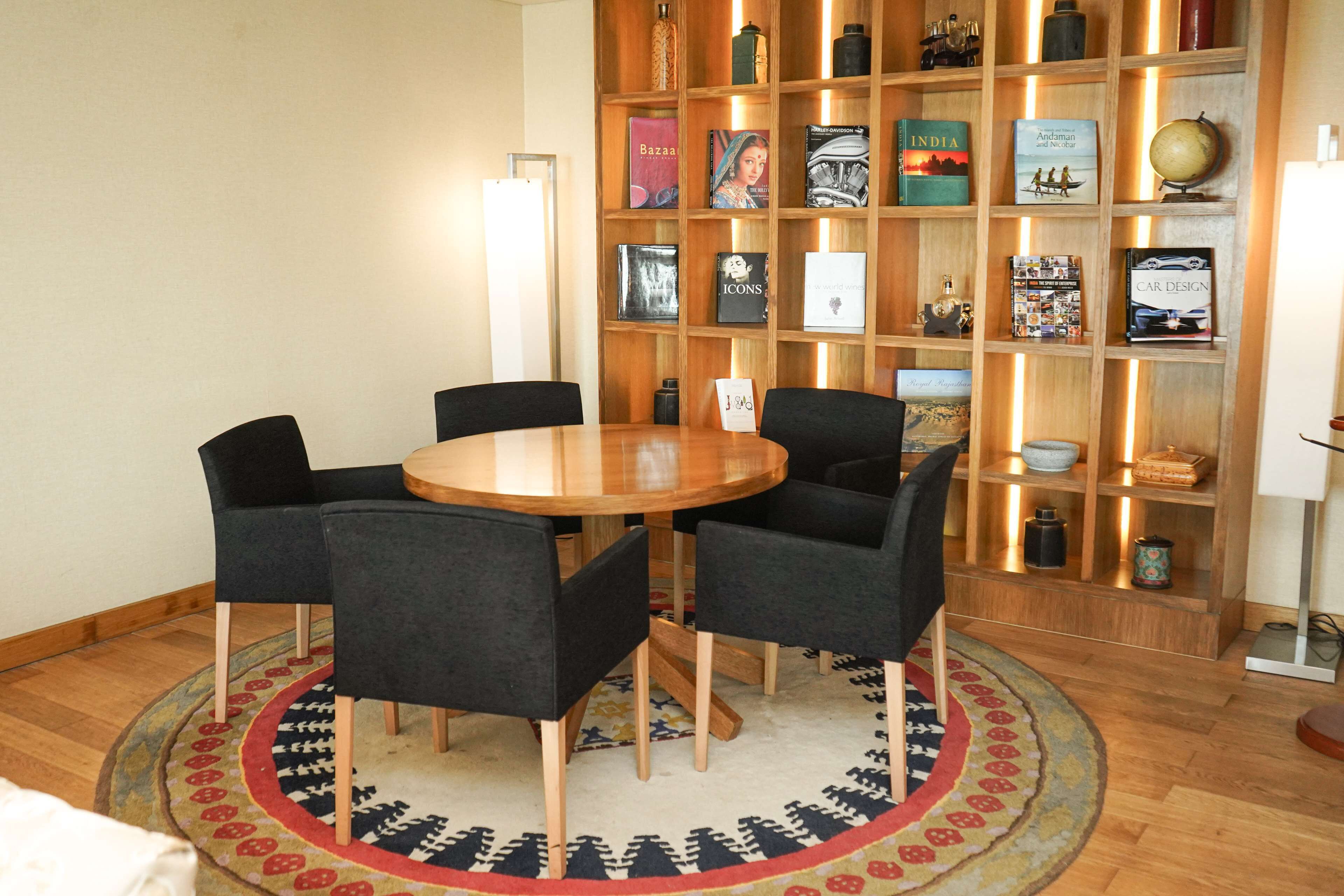 Een kamer met een ronde houten tafel, omringd door vier stoelen, een boekenplank met boeken en decoratieve objecten, en een kamerkleed met een patroon op de vloer.