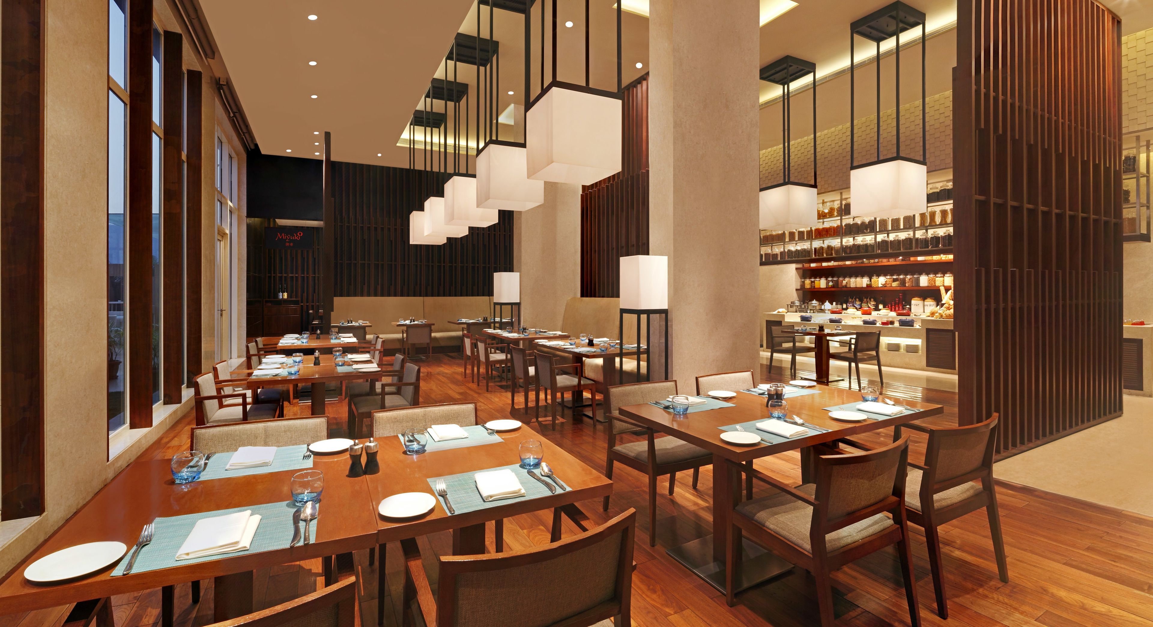 Un restaurant moderne avec des tables et des chaises en bois, des luminaires suspendus et un bar doté d’étagères remplies de bouteilles.