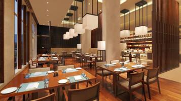 A modern restaurant with wooden tables and chairs, hanging pendant lights, and a bar area with shelves of bottles.