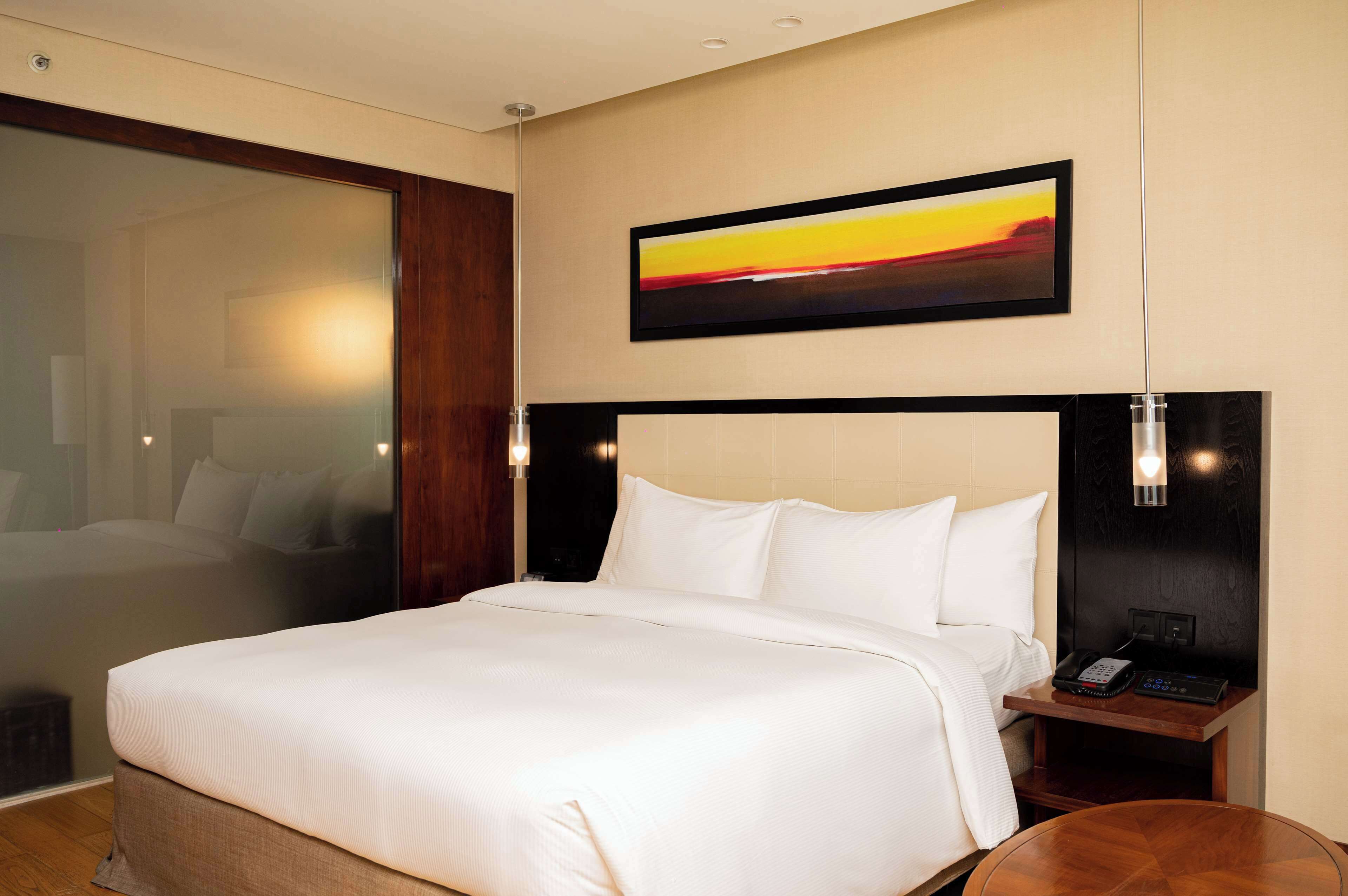 A hotel room with a large bed, a bedside table with a phone and a lamp, a painting on the wall, and a view through a glass door.