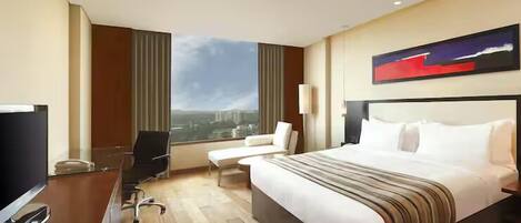 A hotel room with a large bed, a desk with a TV, a chair, a window with a city view, and a painting on the wall.