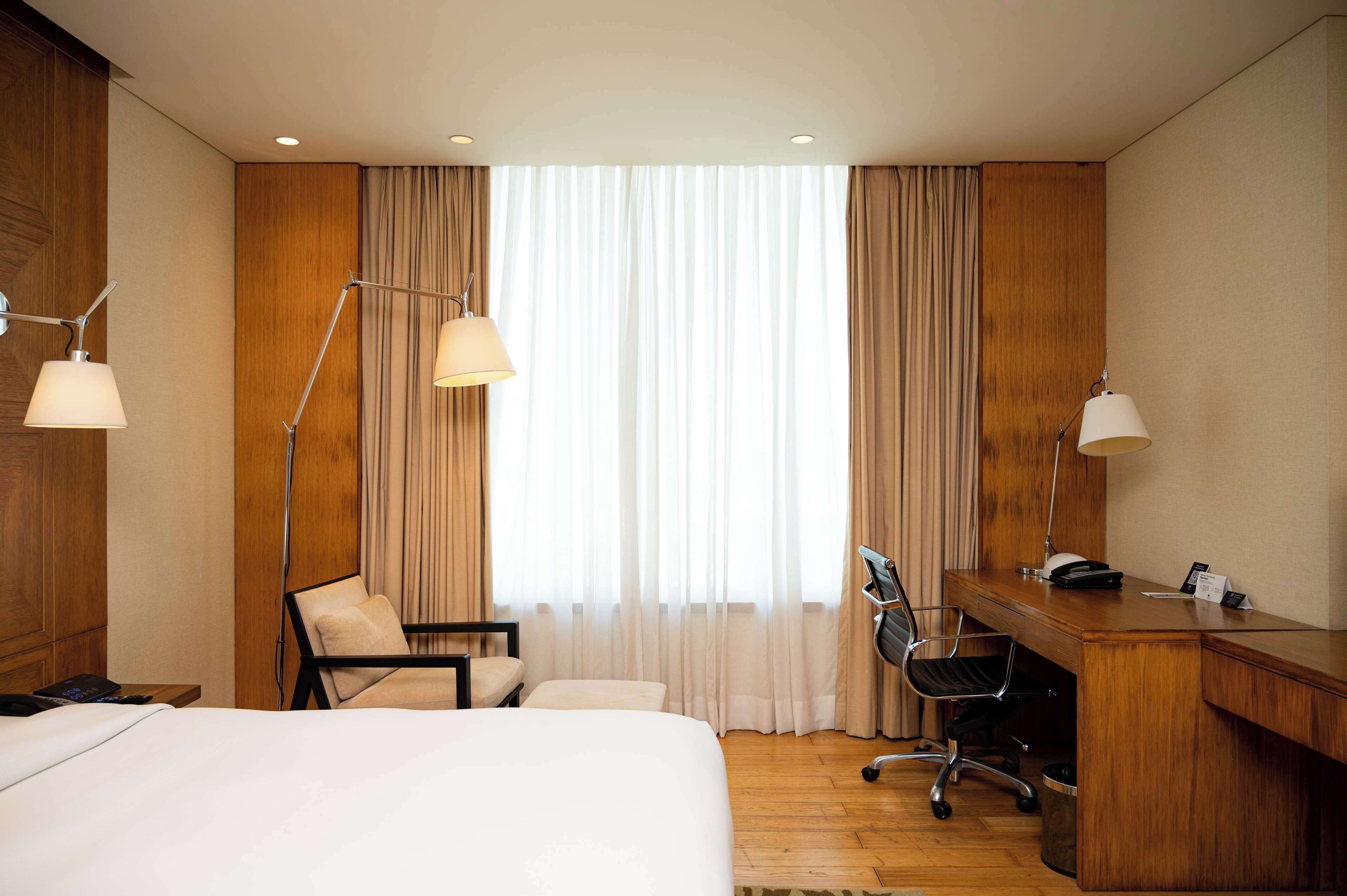 A hotel room with a bed, desk, chair, and large window with curtains.