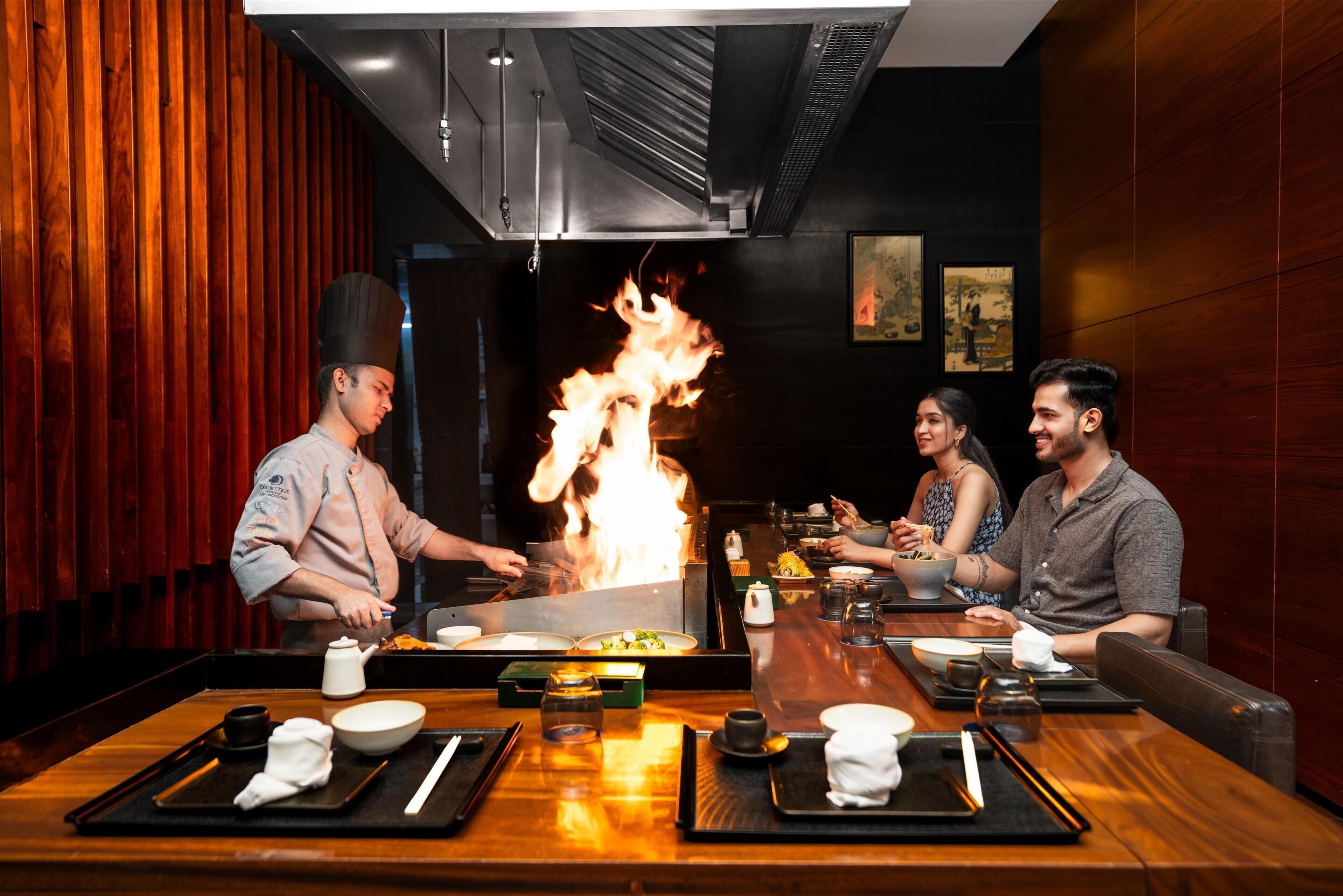 A restaurant with a central cooking area featuring a large fire, wooden walls, and a modern interior design.