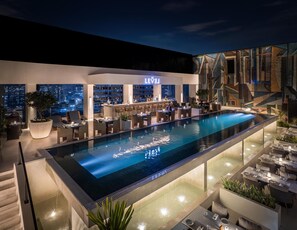 Un área de piscina en la azotea con bar y asientos, con vistas a un paisaje urbano nocturno.